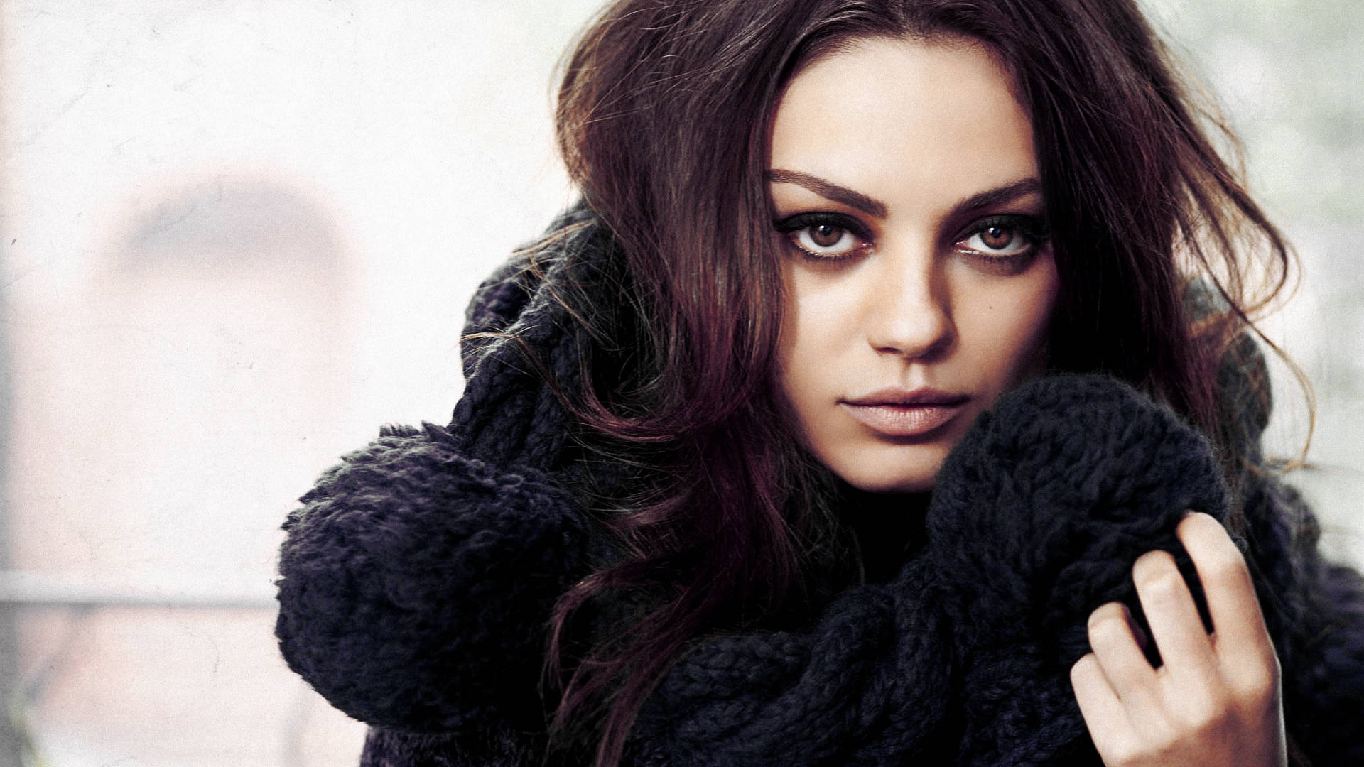 好萊塢神秘美人Mila Kunis 壁紙 — 一位深色頭髮、眼神銳利的女性，在模糊的城市背景下穿著一件厚實的黑色針織外套。