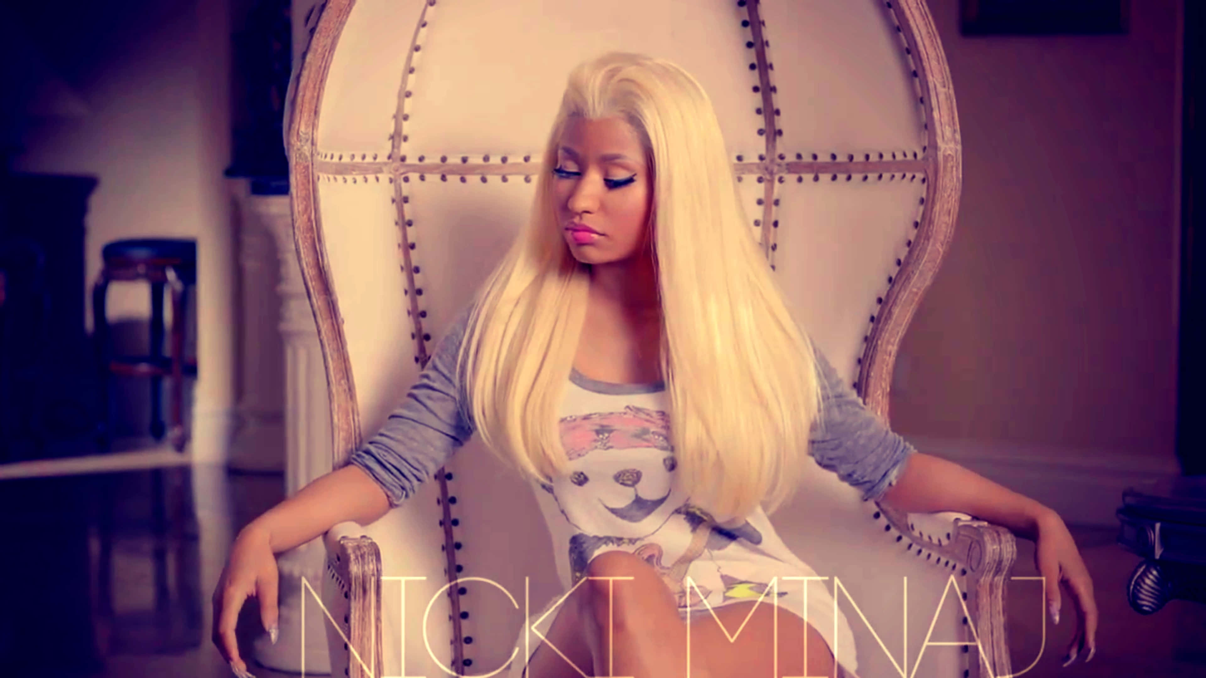 妮琪·米娜 桌布 壁紙 — 妮琪·米娜（Nicki Minaj）端莊地坐在一張華麗的大椅子上，長長的金髮和圖案T恤，房間光線柔和。