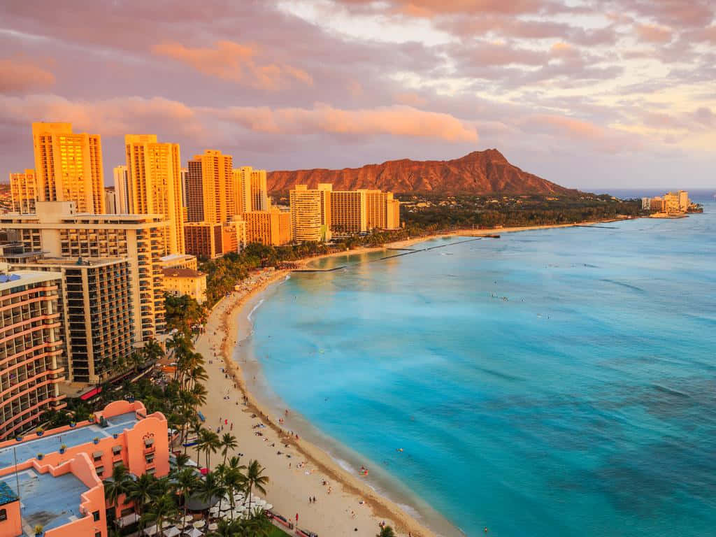 Hawaiian Island 桌布 壁紙 — 威基基海灘日落時的鳥瞰圖，金色摩天大樓、棕櫚樹和鑽石頭山在溫暖的粉紅色和橙色雲彩下閃耀。
