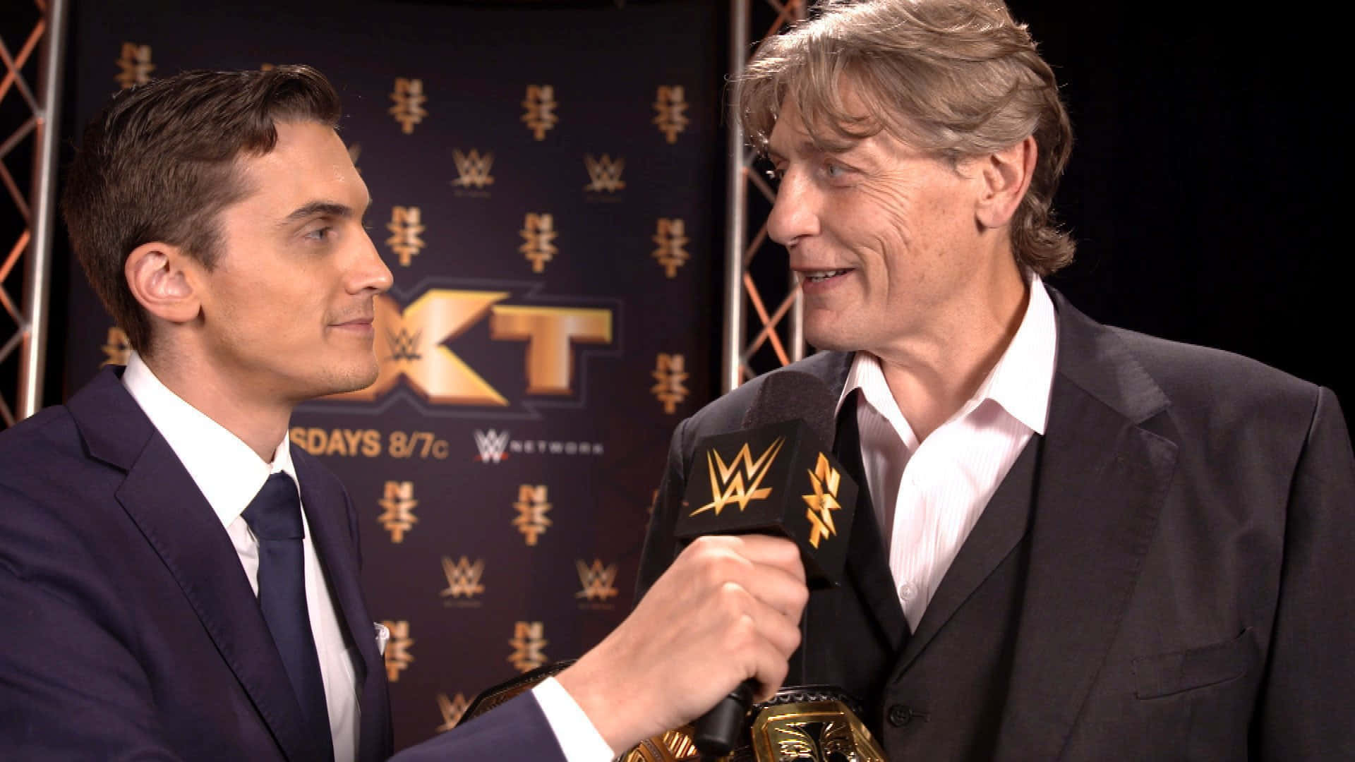 威廉·雷加爾 WWE NXT採訪照片 壁紙 — 兩名身穿西裝的男子在WWE NXT訪問中面對面，背景有冠軍腰帶和品牌標誌。