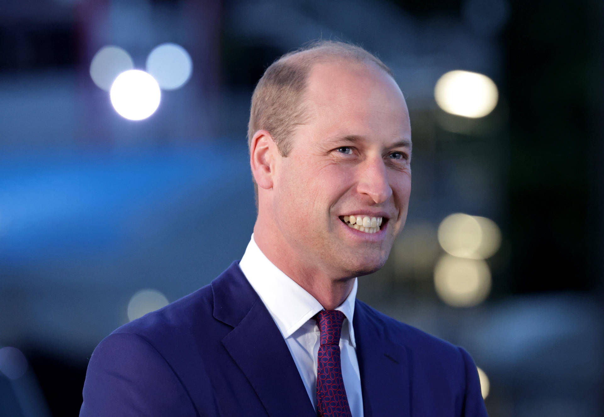 威廉王子身穿海軍藍西裝和圖案領帶，在晚間活動中微笑。