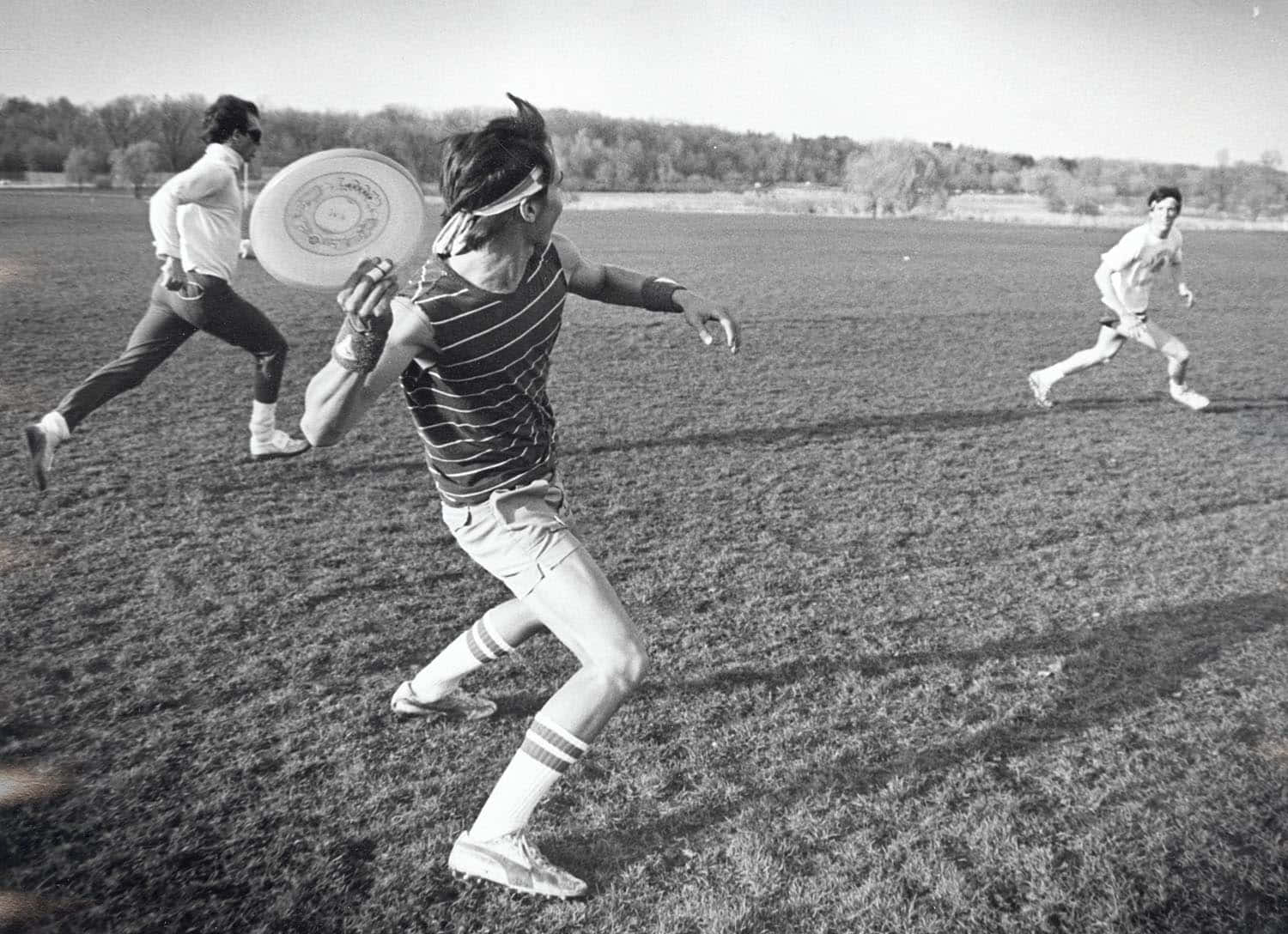 威斯康辛大學最佳 Ultimate Frisbee 背景 壁紙 — 三人在陽光明媚的草地上玩飛盤，背景有樹木。