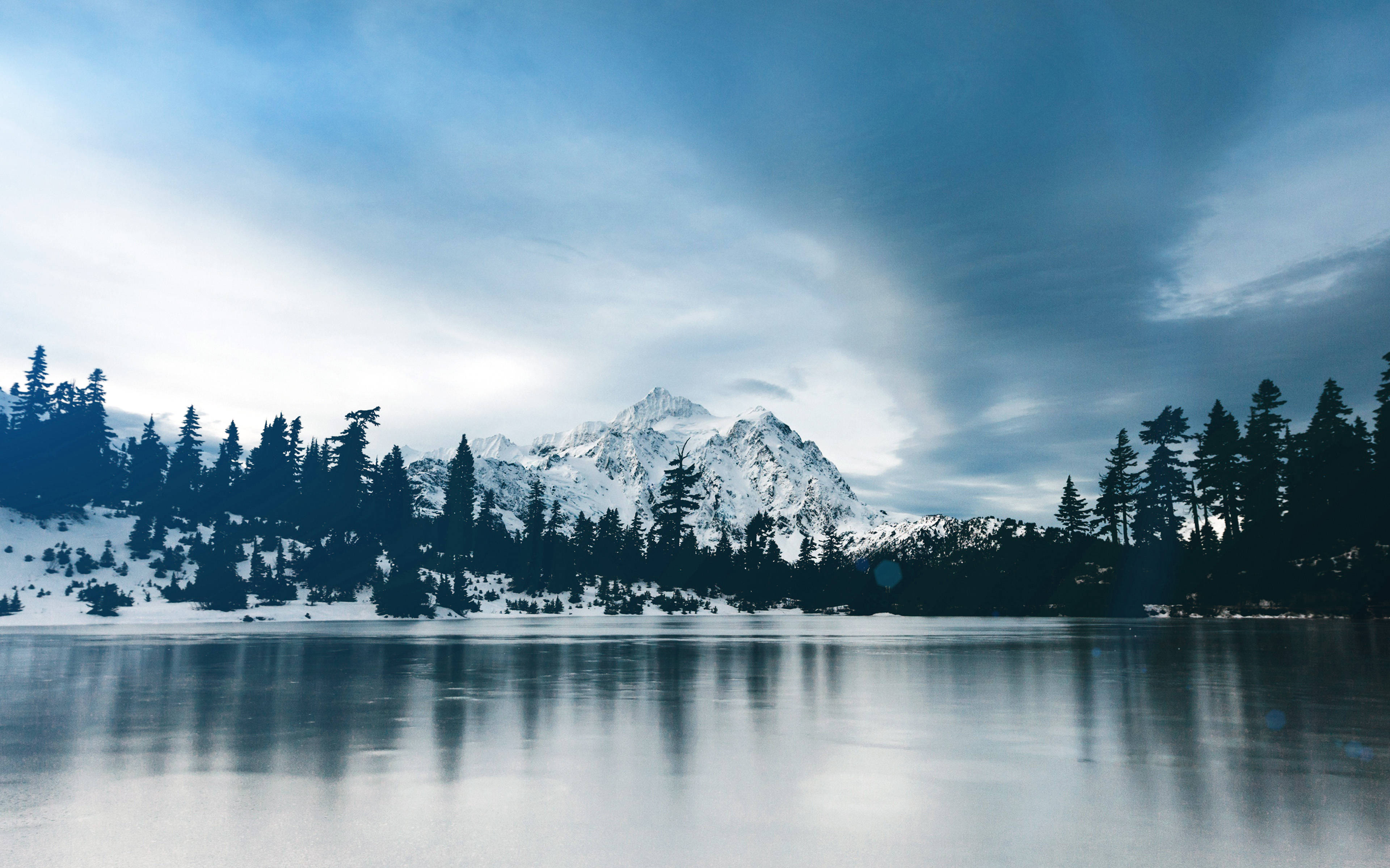 威斯康辛州森林湖泊 壁紙 — 寧靜的冬日湖面倒映著積雪的山峰，天空憂鬱的藍色中飄著縷縷薄雲。