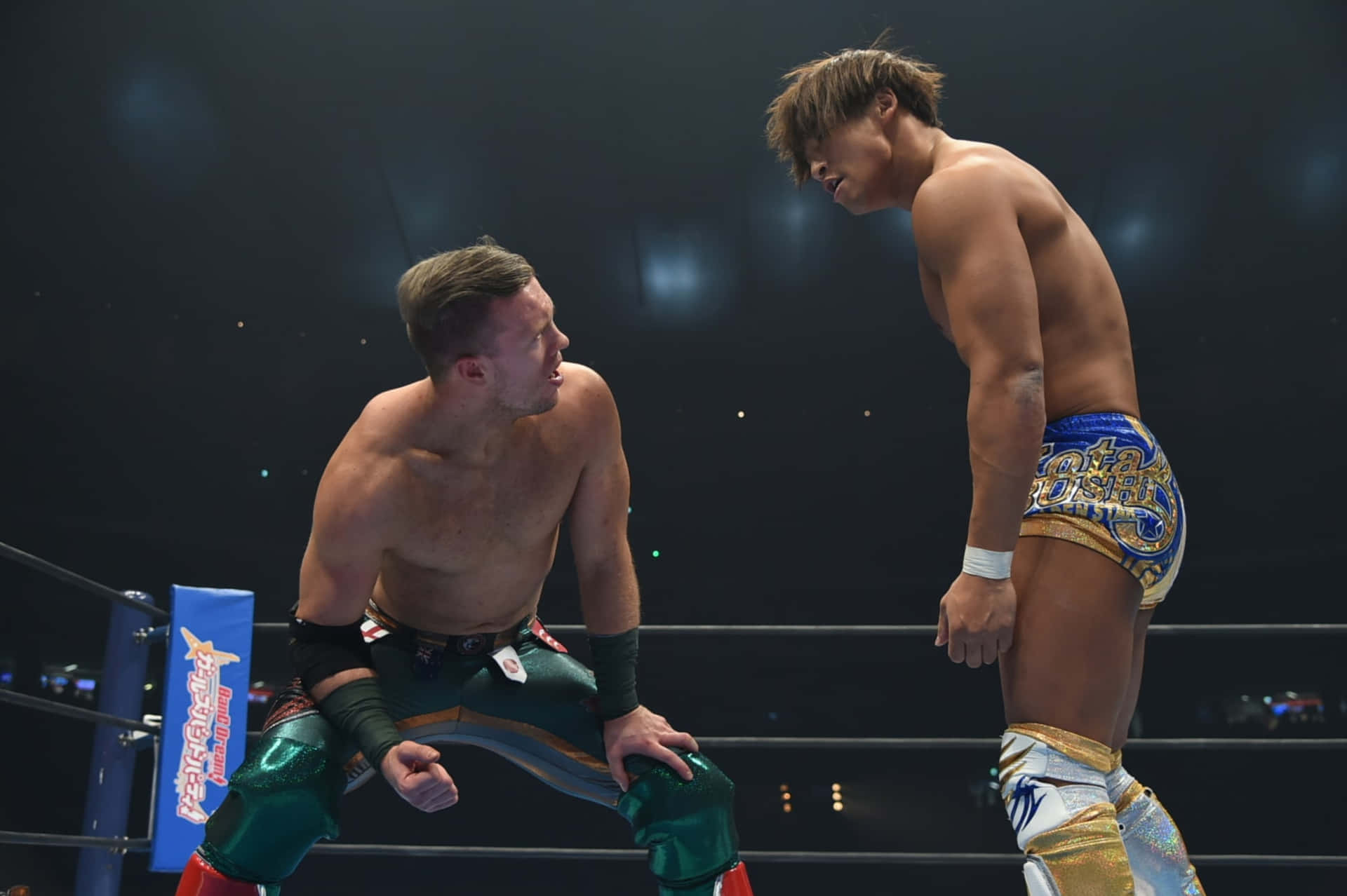 威爾·奧斯普雷與 Kota Ibushi 壁紙 — 兩名肌肉發達的摔角手在明亮的擂台上，於場館燈光下激烈對峙。