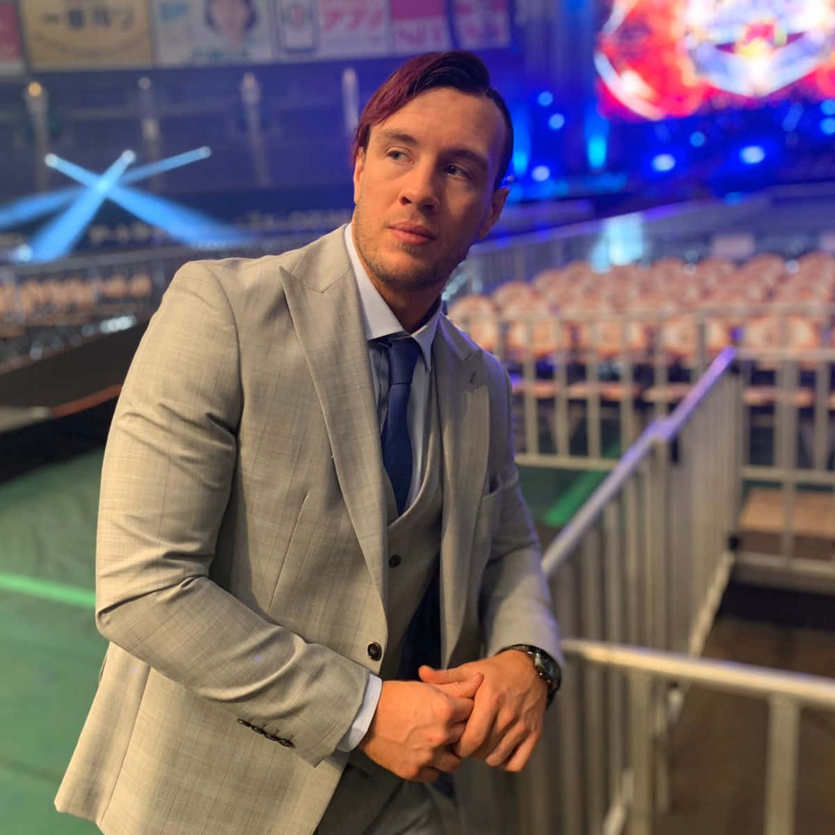 灰色西裝男子靠在競技場欄杆上，身後有彩色燈光和空座位。