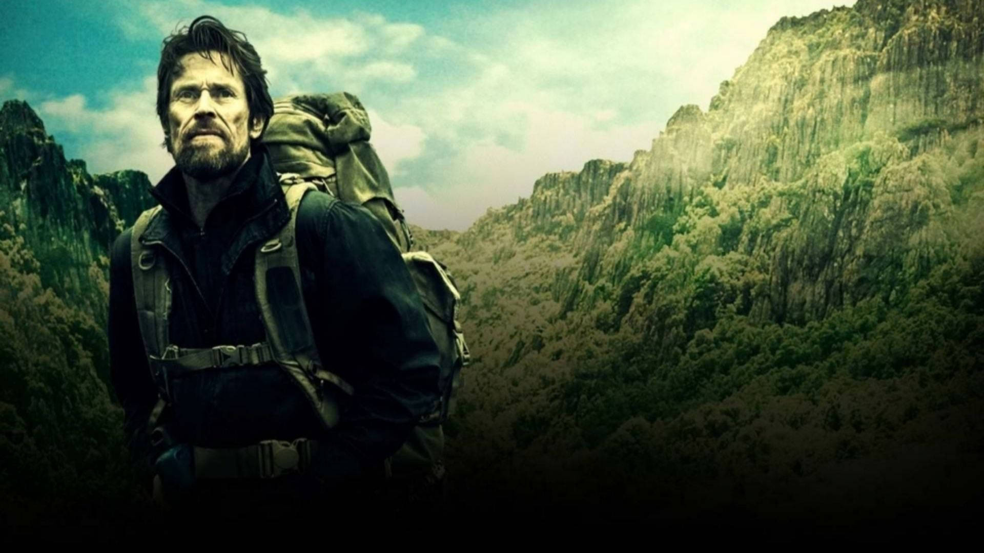 威爾·達福登山冒險 壁紙 — 一個粗獷的鬍鬚男背著一個大背包，在陰天背景下，以高聳的綠色山脈為背景，莊嚴地站立。