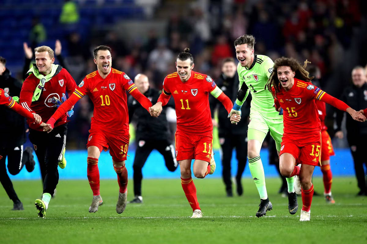 Football Players 桌布 壁紙 — 威爾斯足球員身穿紅色球衣在綠色球場上奔跑，慶祝入球，狂喜並手臂相連。