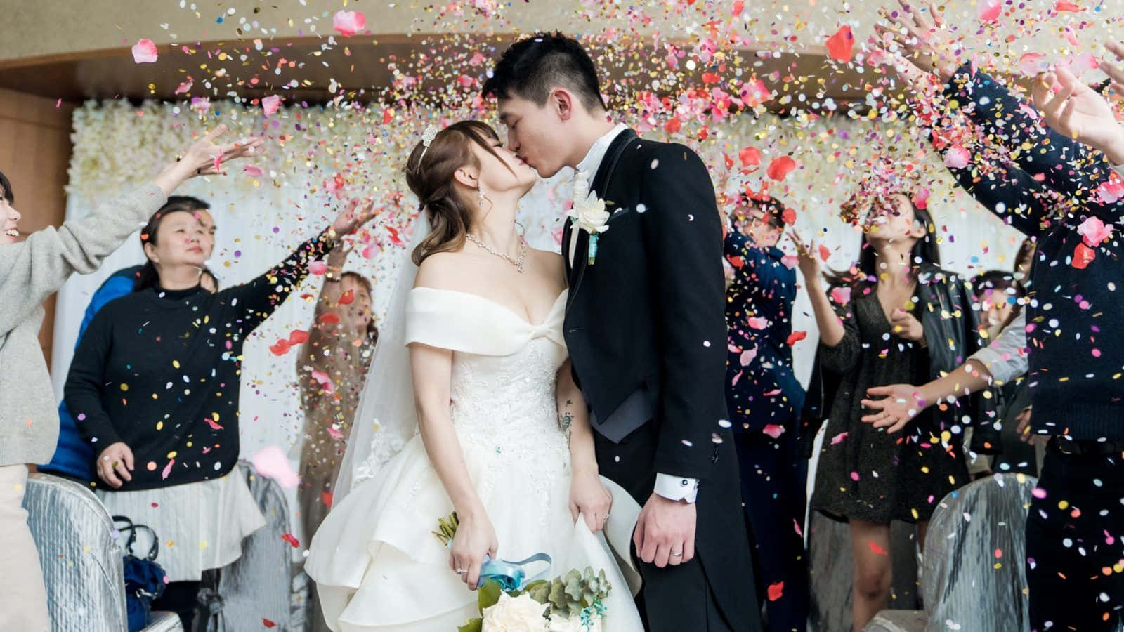 婚宴派對親吻照片 壁紙 — 新娘和新郎在五彩紙屑的洗禮下接吻，賓客在他們周圍慶祝。