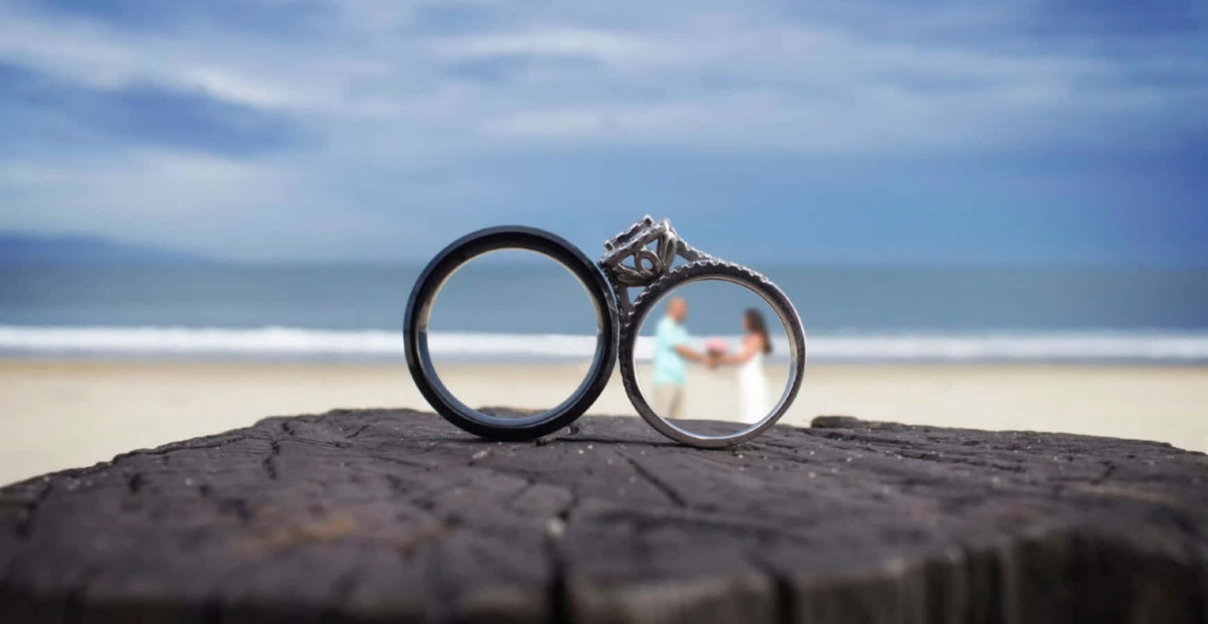 兩枚結婚戒指放在木頭上，身後沙灘上有一對情侶牽手。