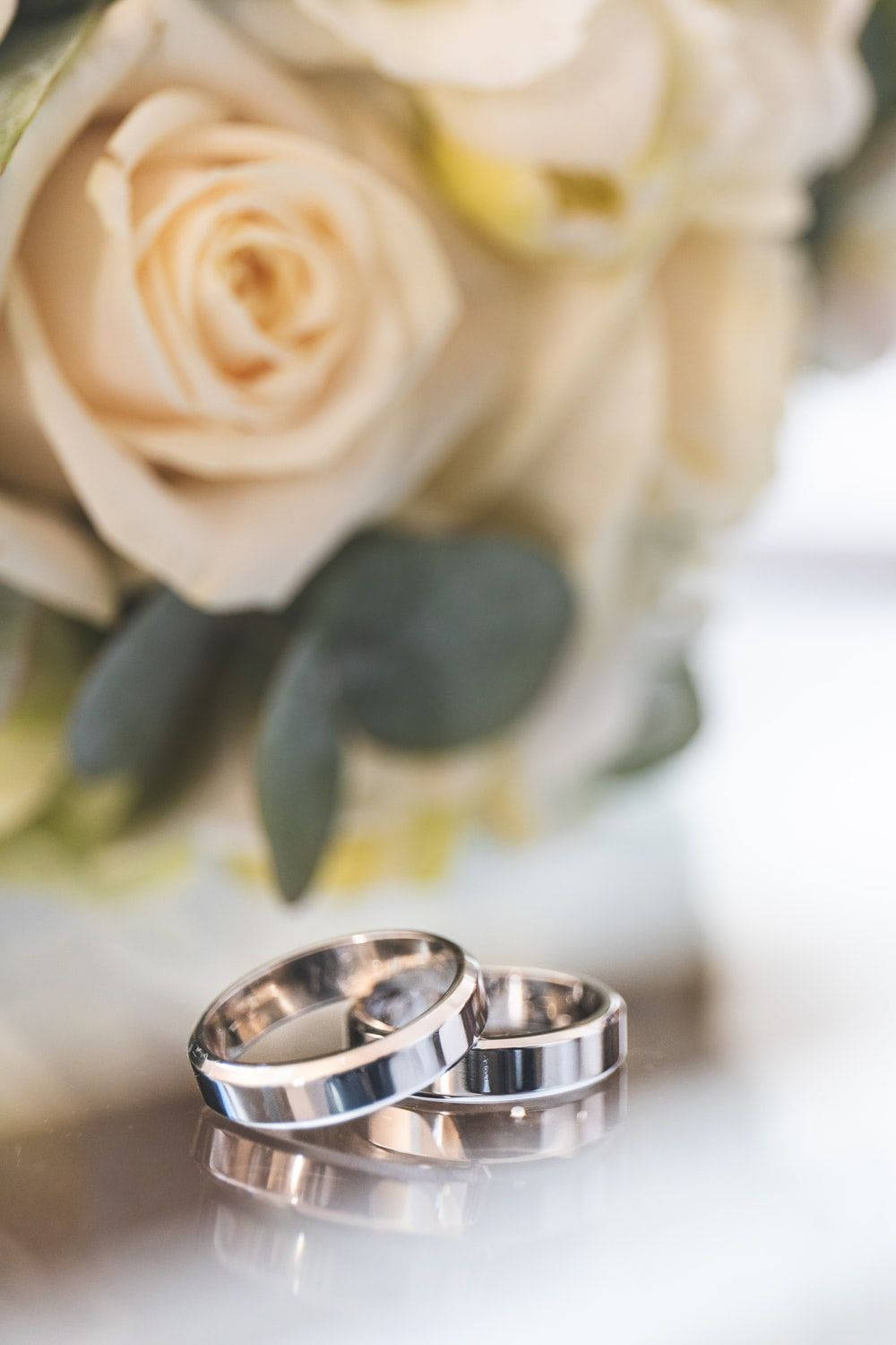 婚戒黃色花束 壁紙 — 兩隻閃亮的結婚戒指放在反光表面上，後面有柔焦的奶油色玫瑰和尤加利葉花束。
