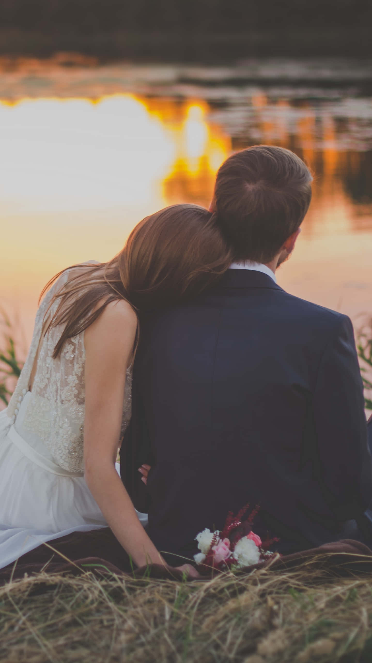 婚禮情侶湖畔休憩 壁紙 — 一對穿著婚紗禮服的新人背對背坐在草地上，凝視著倒映在水面上的金色夕陽。