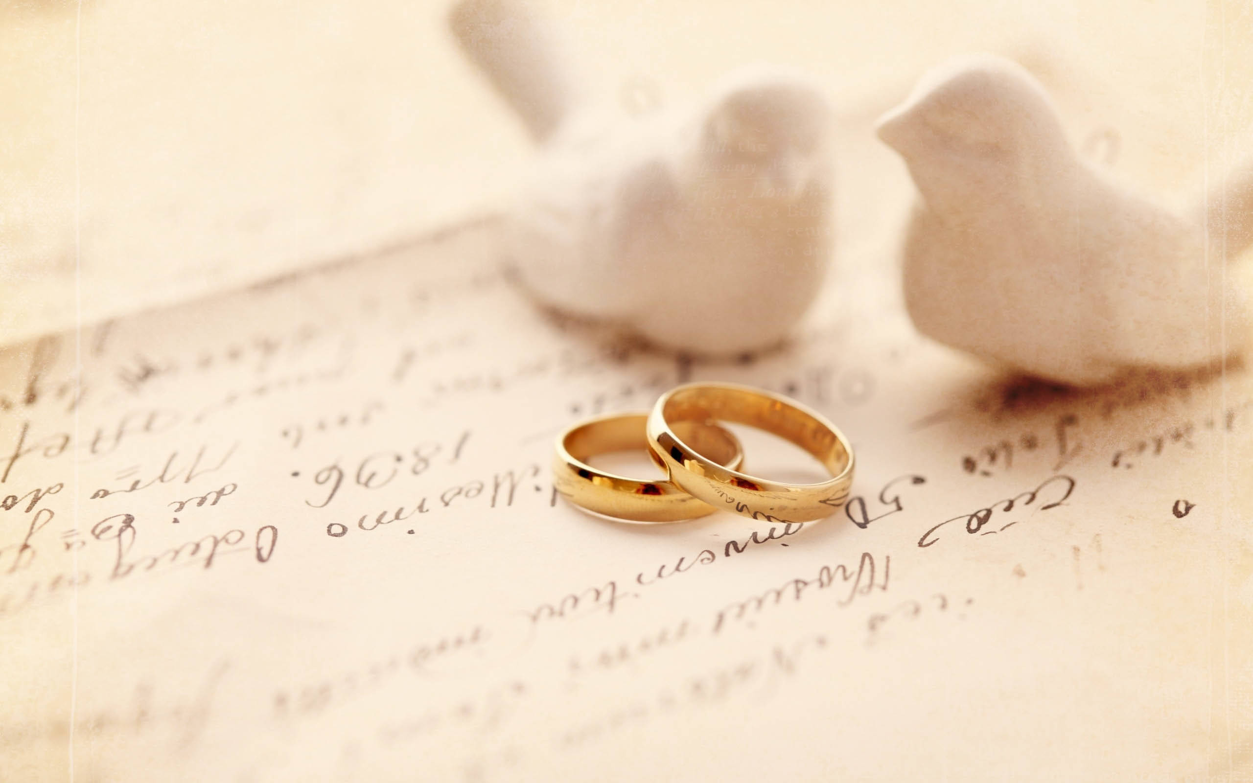 婚禮情鳥 壁紙 — 兩枚金色結婚戒指放在復古手寫紙上，旁邊有白色陶瓷鳥，在柔和溫暖的光線中。