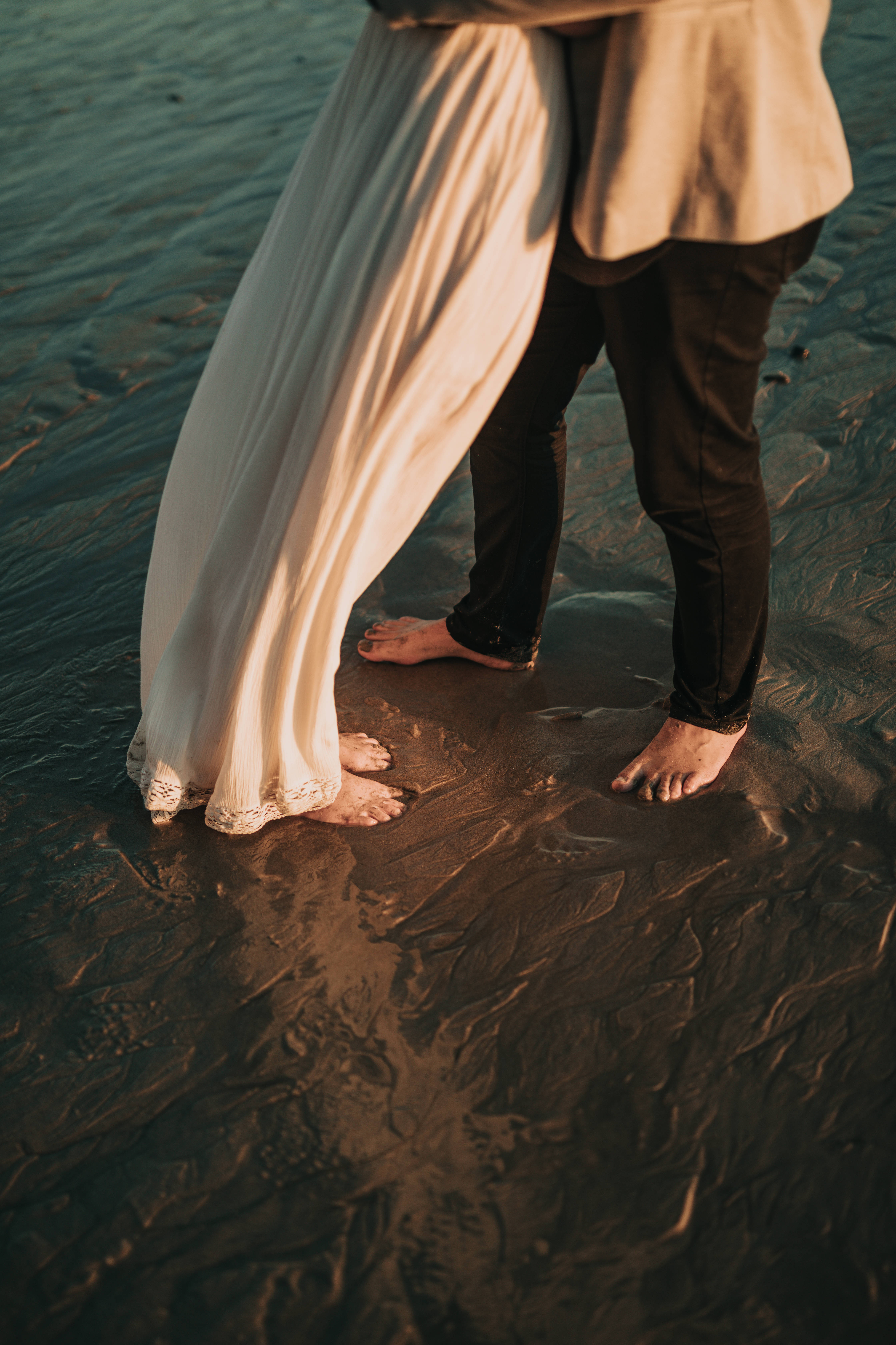婚禮美學海灘 壁紙 — 一對情侶赤腳站在夕陽下的淺水中，雙腳觸碰，布料在他們周圍飄動。