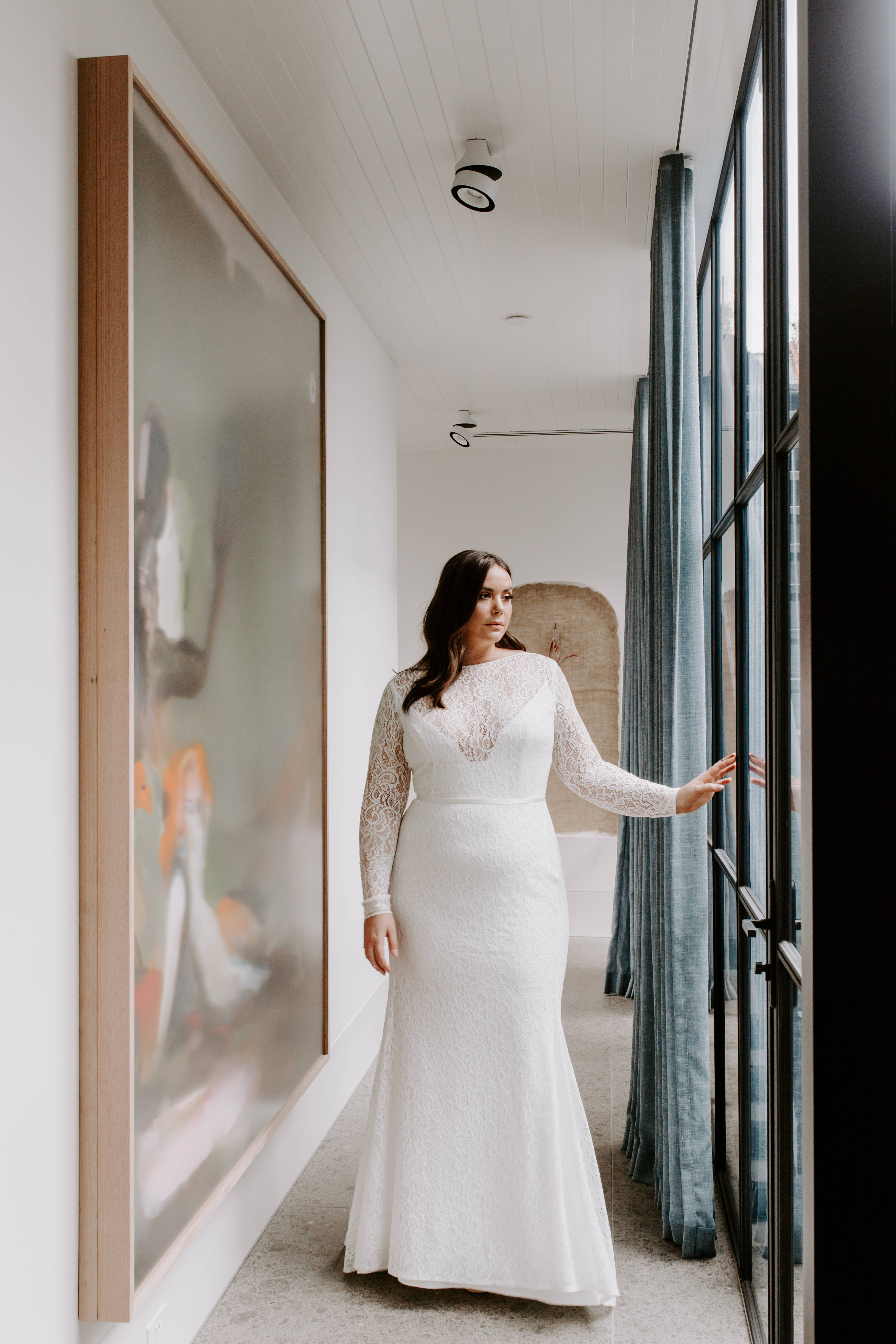 婚禮風格新娘窗邊 壁紙 — 一位身穿白色蕾絲婚紗的女性站在現代走廊中，觸摸著大型抽象畫旁邊的玻璃門。