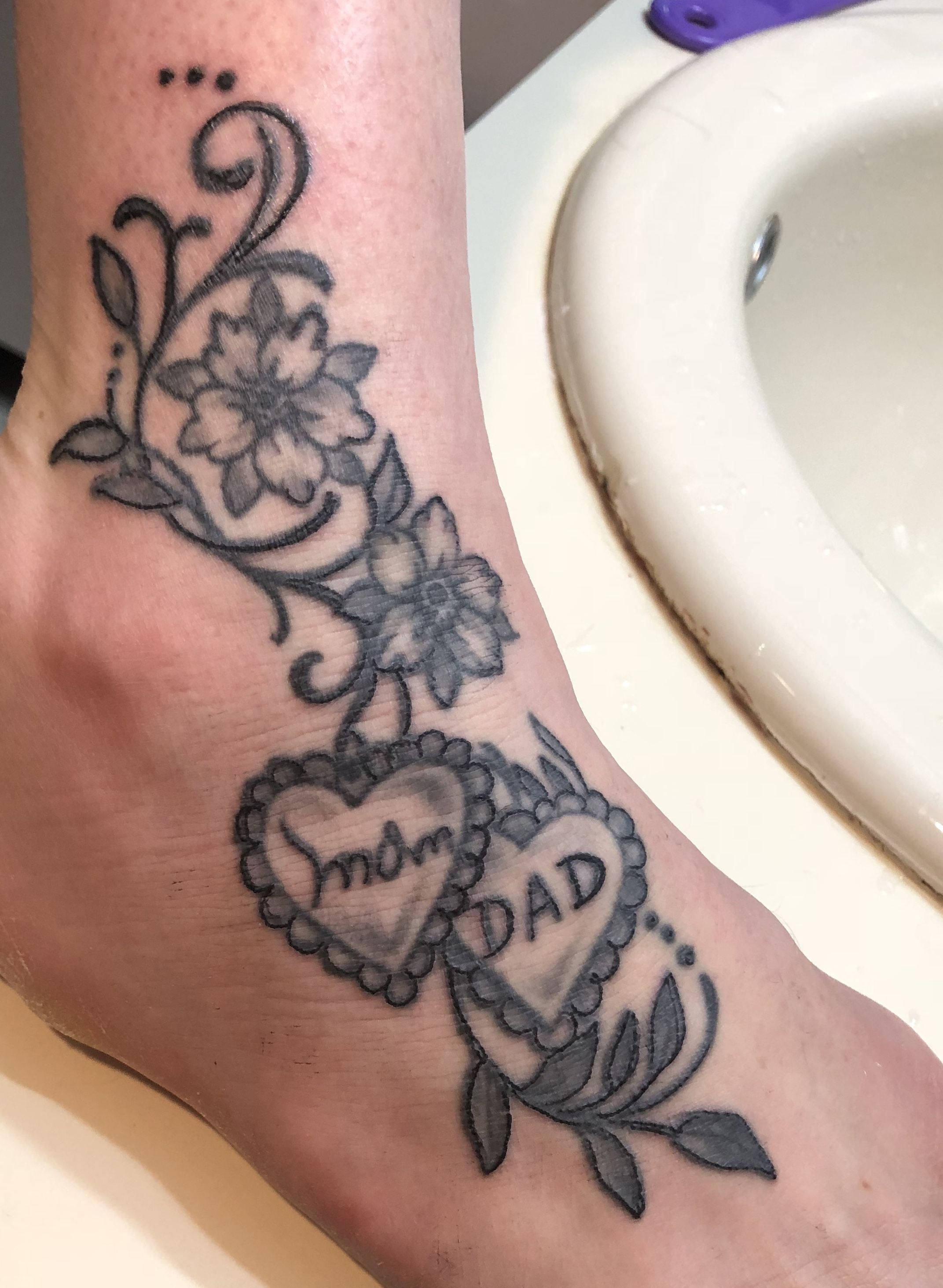 媽媽與爸爸 腳踝紋身 壁紙 — 腳踝上的黑灰色紋身，有花卉藤蔓和兩個心形吊飾，上面刻有Mom和Dad。
