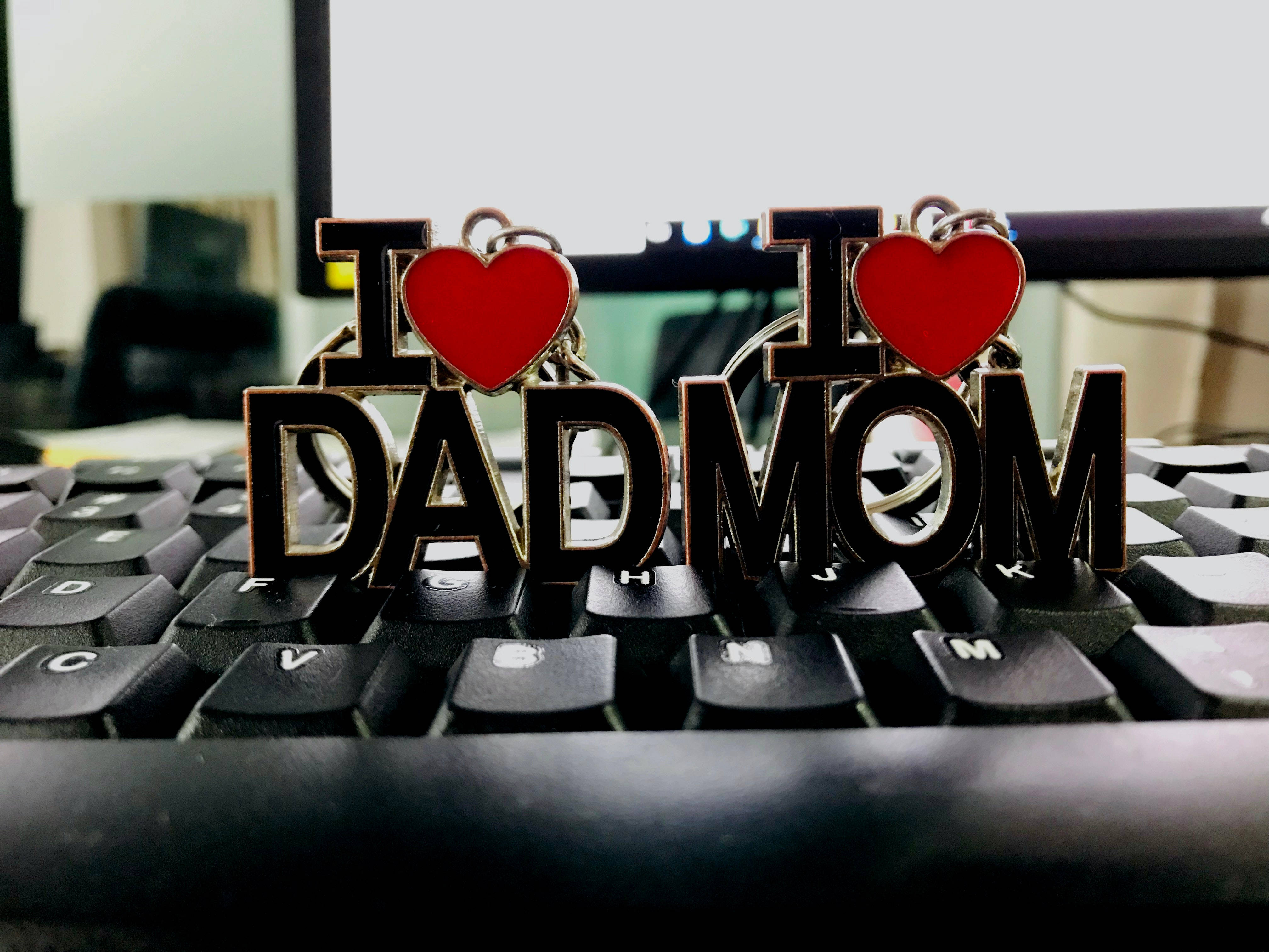 媽媽與爸爸 鑰匙圈 壁紙 — 兩個金屬鑰匙扣，分別拼寫 I heart DAD 和 I heart MOM，放在一個黑色電腦鍵盤上，背景是模糊的辦公室。