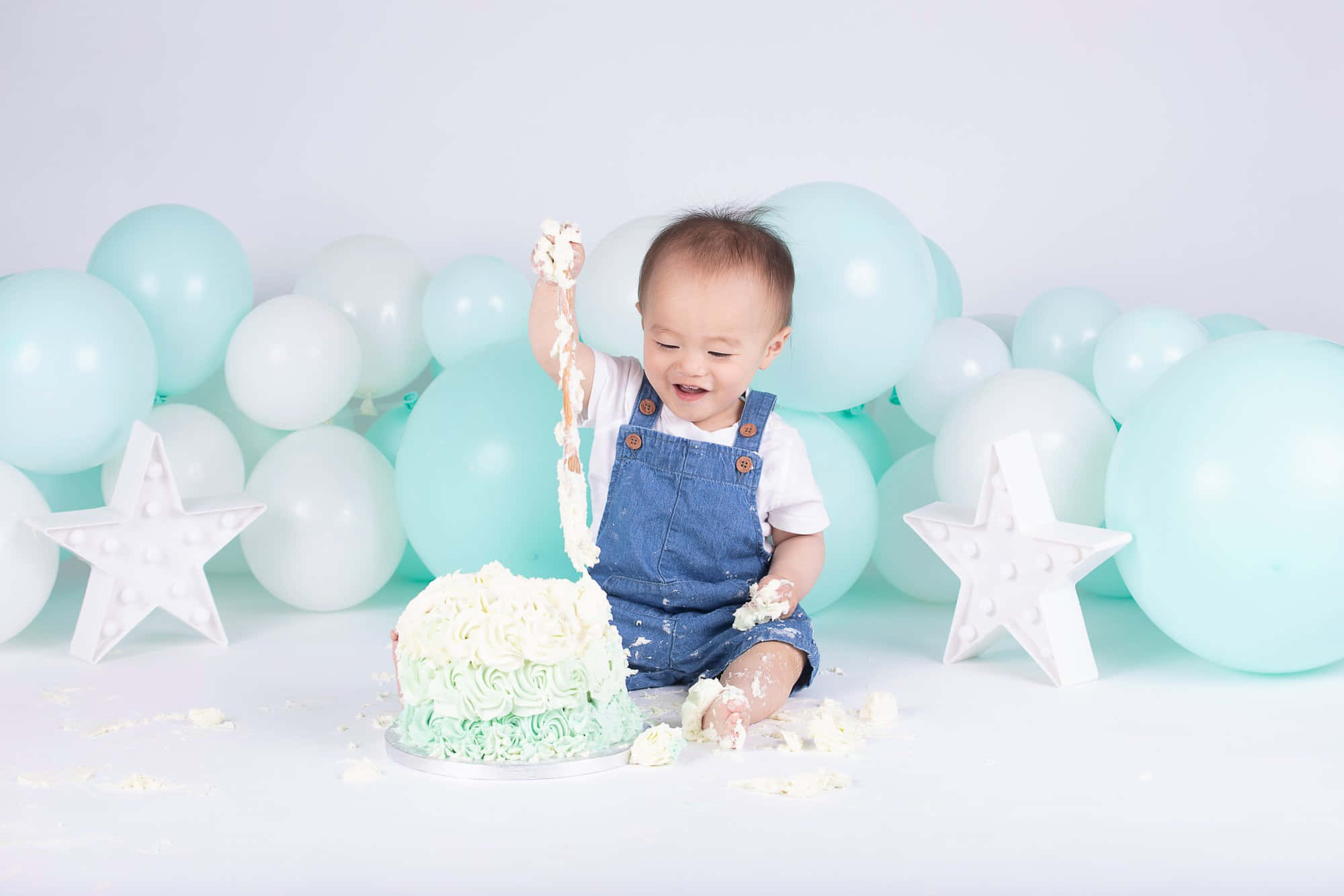 嬰兒生日派對的柔和蛋糕砸臉照片 壁紙 — 一個快樂的嬰兒穿著牛仔工人褲，熱情地砸碎一個薄荷綠蛋糕，糖霜四濺，周圍環繞著粉彩氣球和星星裝飾。