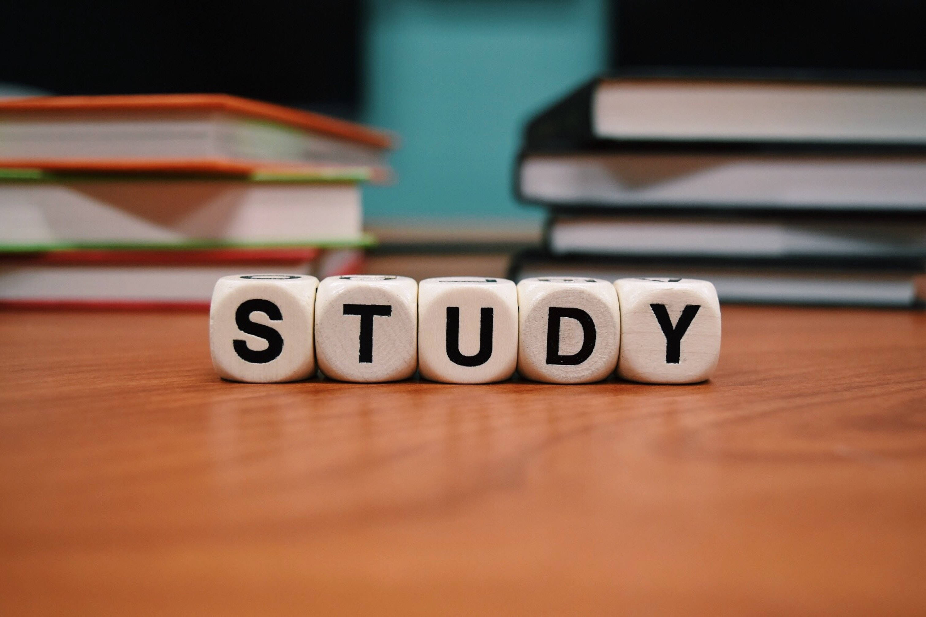 字母骰子學習 壁紙 — 拼出 STUDY 的木製方塊放在書桌上，周圍環繞著成堆的書籍，營造專注的學習環境。