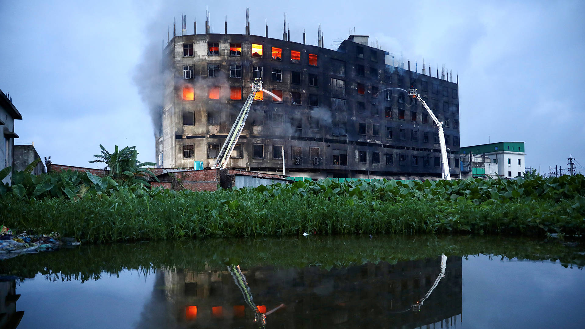 孟加拉工廠火災肆虐 壁紙 — 一座燃燒的多層建築，窗戶發光並冒煙，倒映在平靜的水道上，周圍環繞著綠色植物。