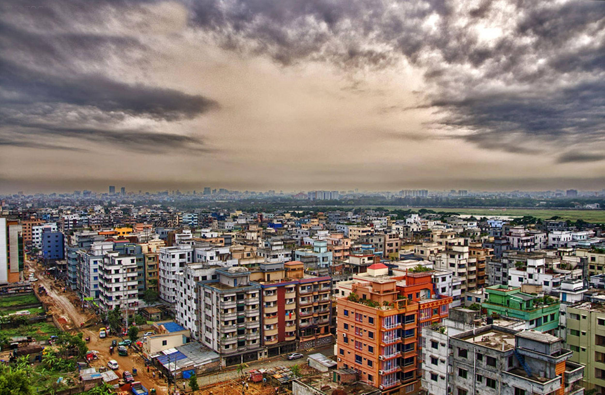 孟加拉達卡的城市全景 壁紙 — 在戲劇性的多雲天空下，廣闊的城市景觀，密集的彩色建築和遙遠的地平線。