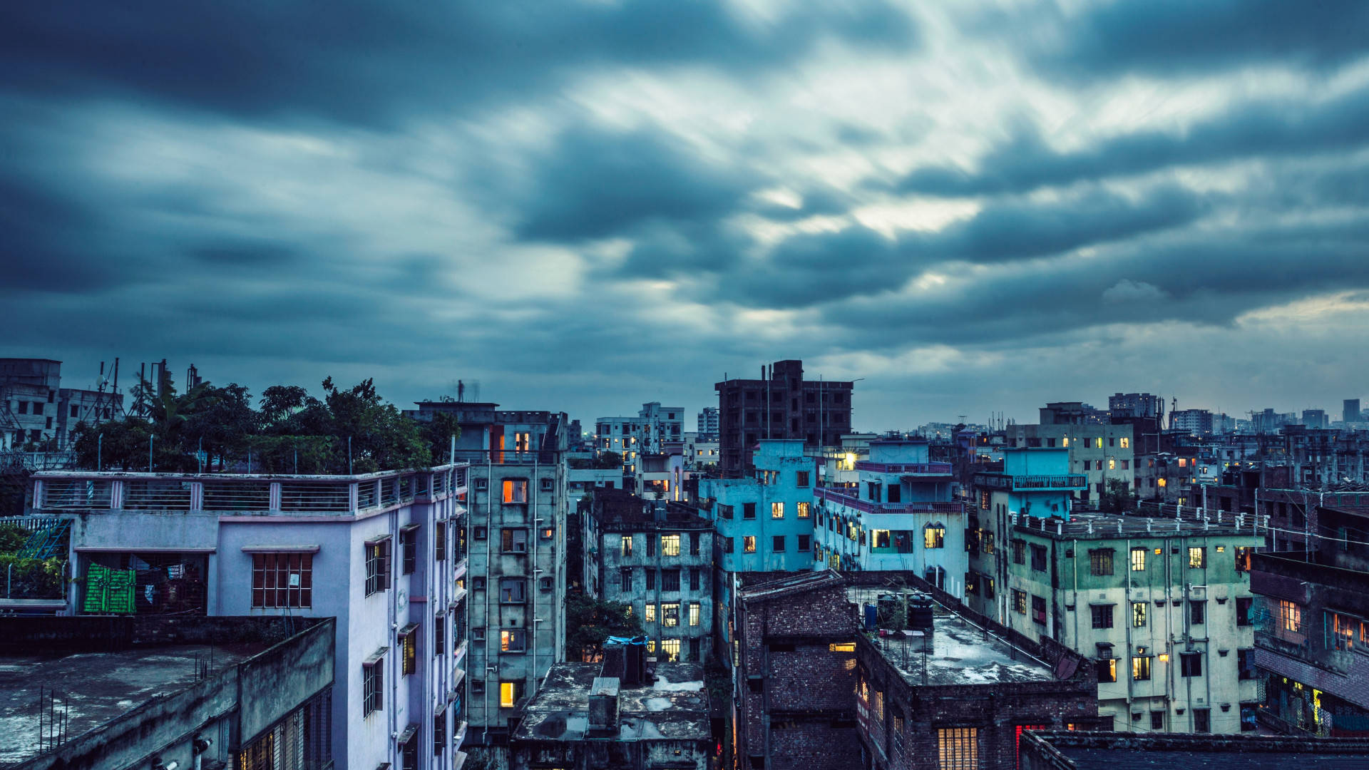孟加拉達卡街區建築 壁紙 — 黃昏時分，陰鬱的城市天際線，帶有條紋雲彩，溫暖的燈光從飽經風霜的建築物窗戶中閃爍。