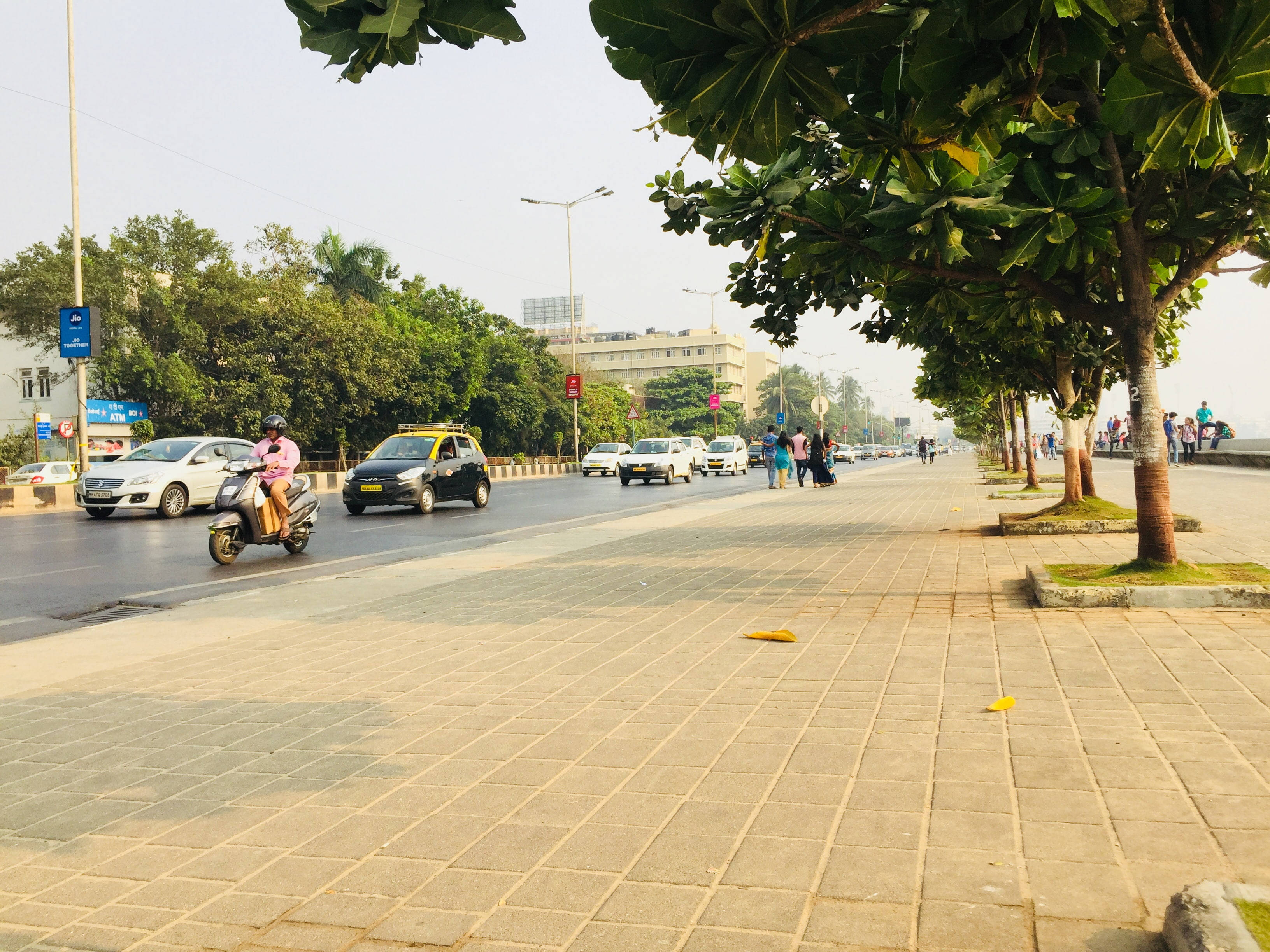 孟買沿海路旁公園 壁紙 — 一條陽光普照的城市街道，行人與車輛在樹蔭下的人行道上移動，天空略帶朦朧。