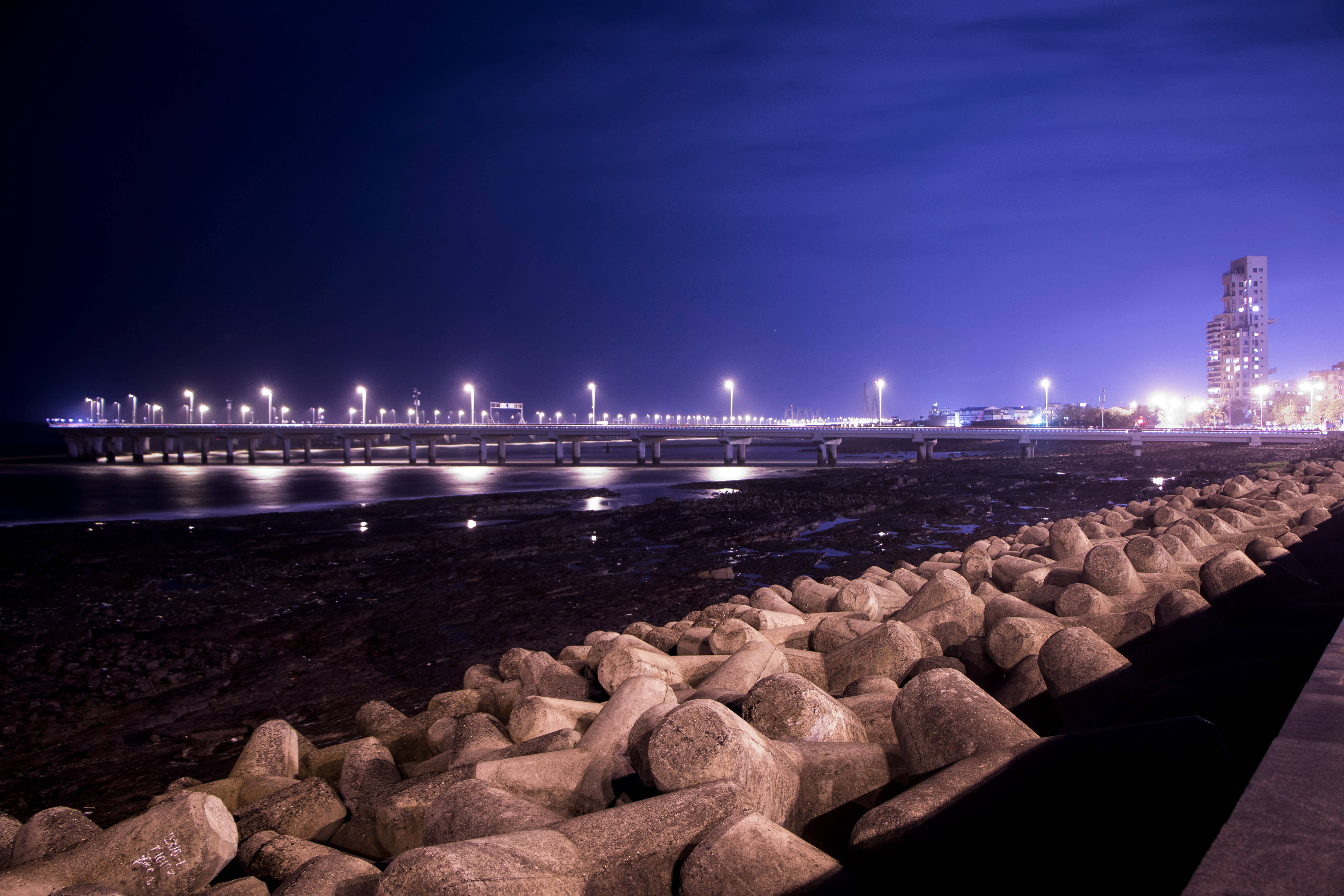 孟買海岸防波堤夜景 壁紙 — 一條長長的照明碼頭在夜間延伸在平靜的水面上，發光的街燈倒映在水面，前景是岩石防波堤。