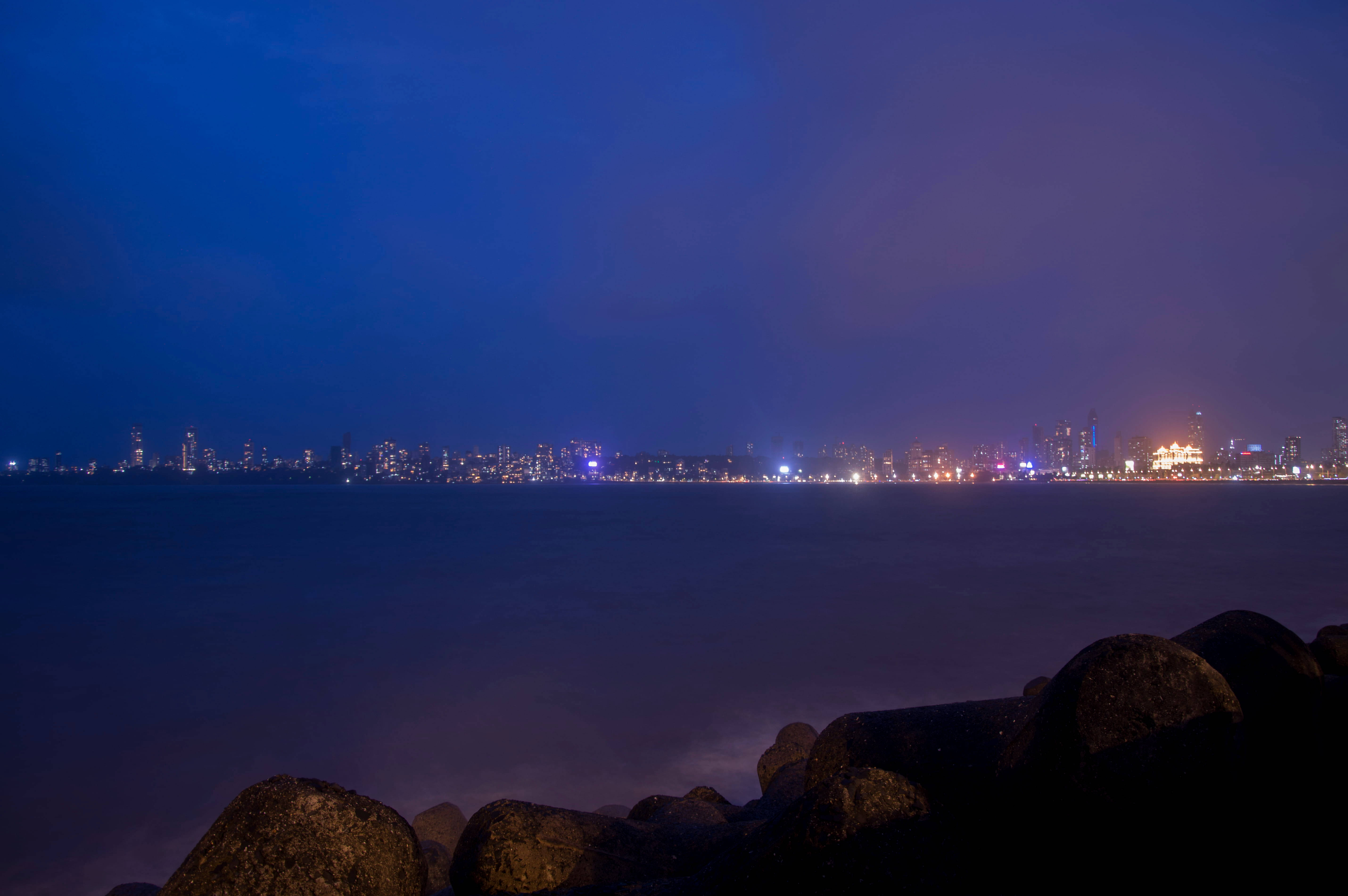 孟買海濱大道城市夜景 壁紙 — 寧靜的夜景，城市天際線在平靜的海面上閃耀，前景有深色岩石。