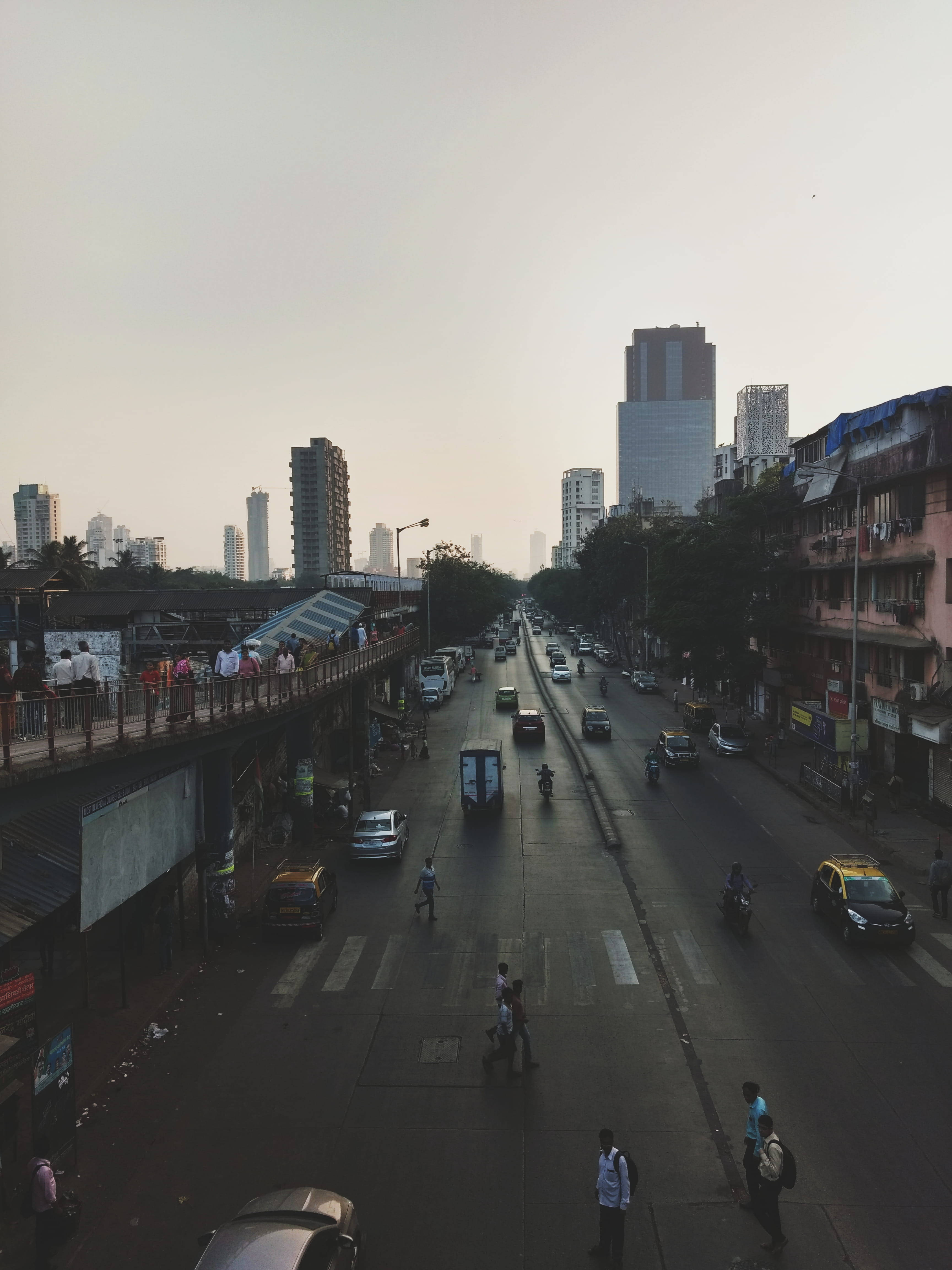 孟買繁忙街景 壁紙 — 黃昏時分繁忙的城市街道，行人過馬路，車輛行駛，高樓大廈在朦朧的天空下聳立。