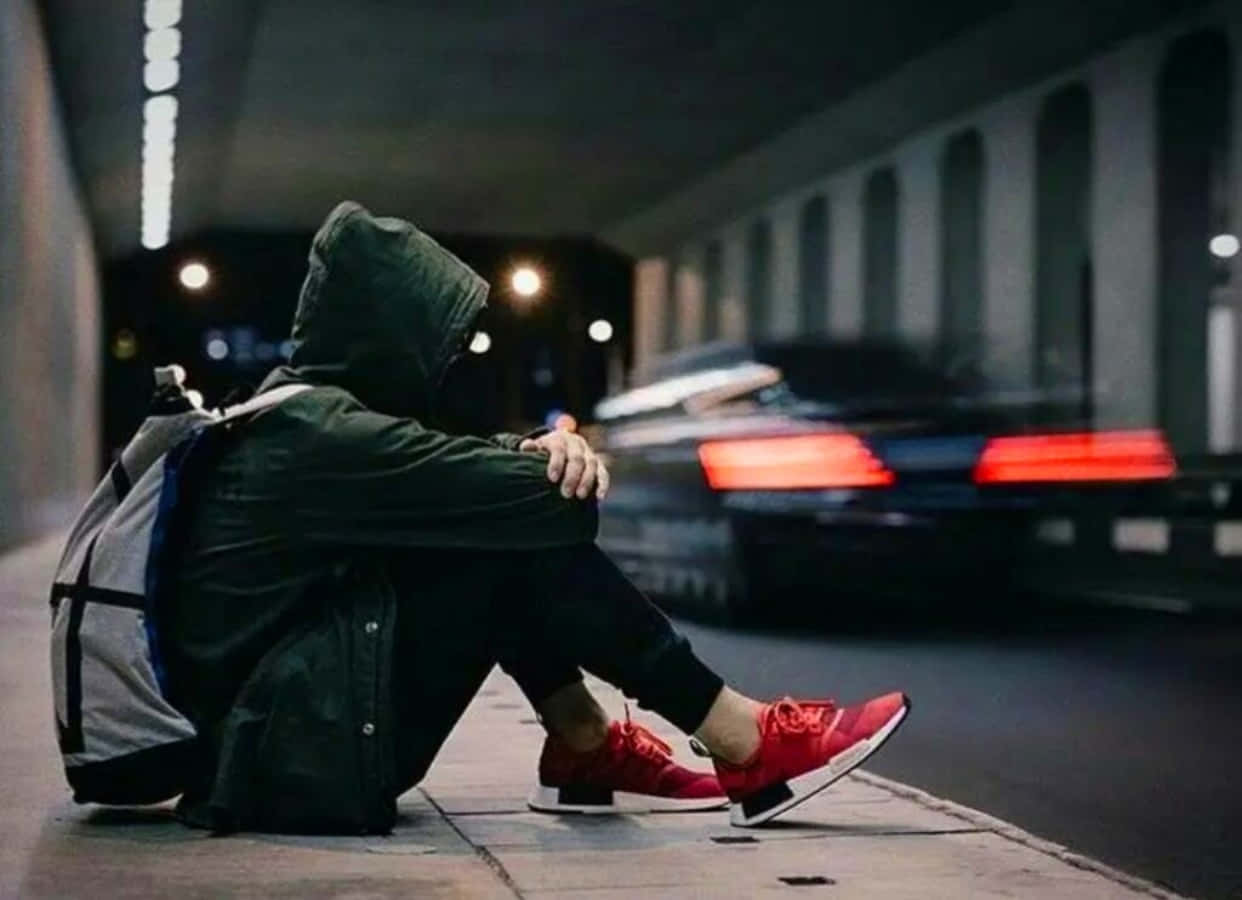 孤獨傷感男子路邊圖 壁紙 — 一個戴著連帽衫的人獨自坐在夜晚的人行道上，一輛汽車帶著紅色尾燈疾馳而過。