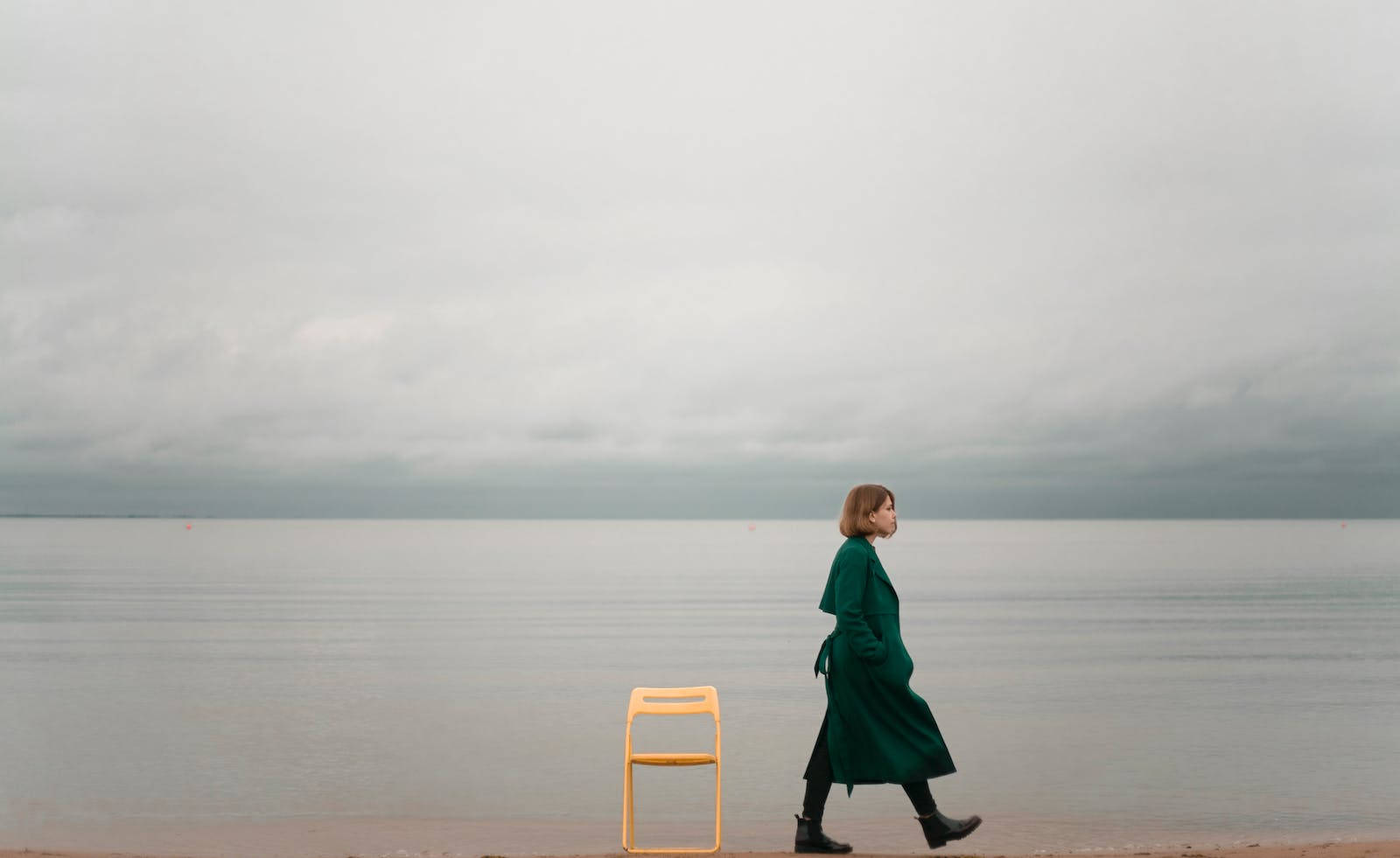 孤獨女子漫步海灘 壁紙 — 一位穿著綠色外套的女性在陰天平靜的海灘上走過一張黃色椅子。