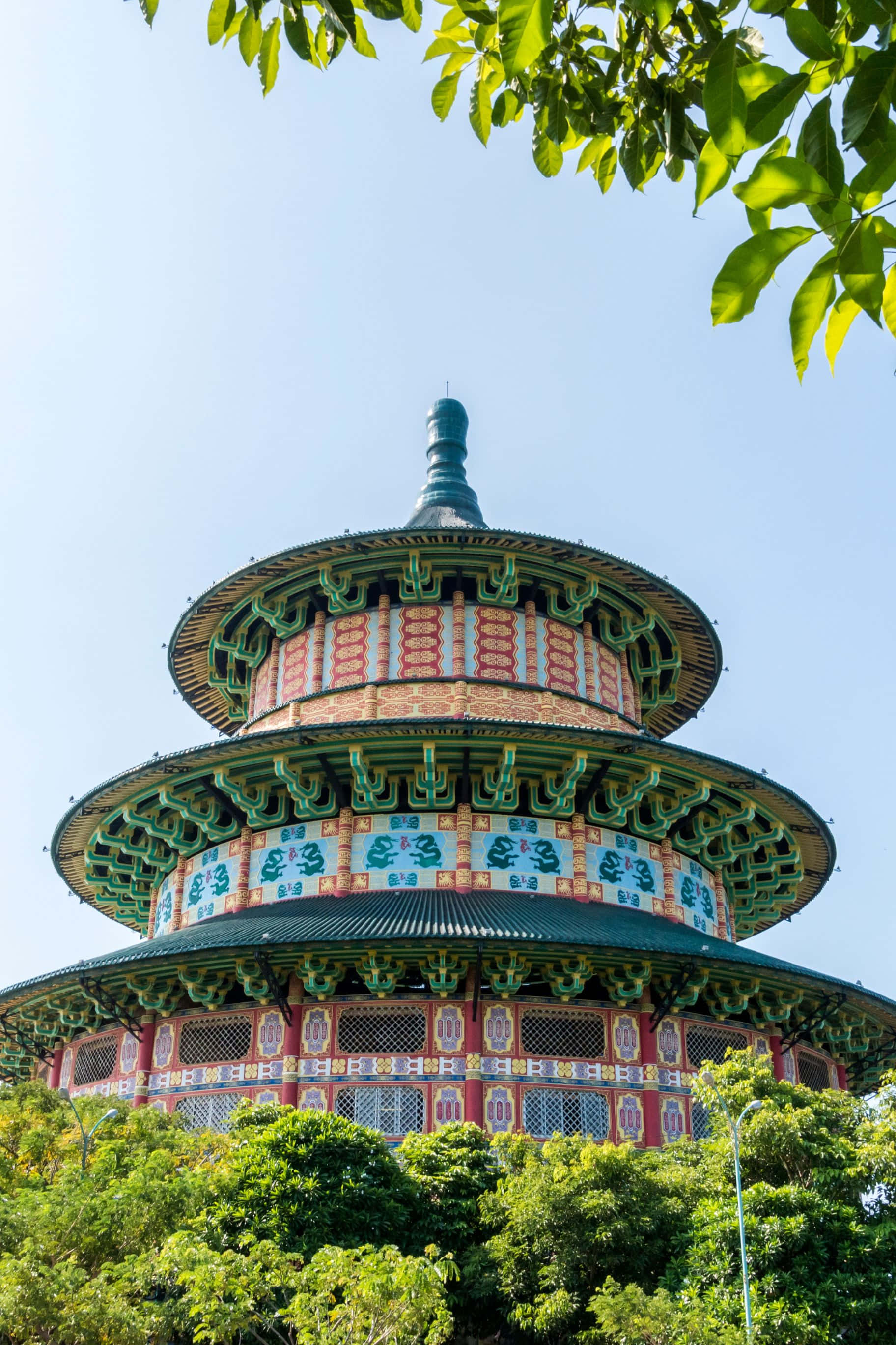 孤獨旅人凝望落日剪影 壁紙 — 一座色彩繽紛的多層傳統寶塔在晴朗的藍天下從茂密的綠葉中升起。
