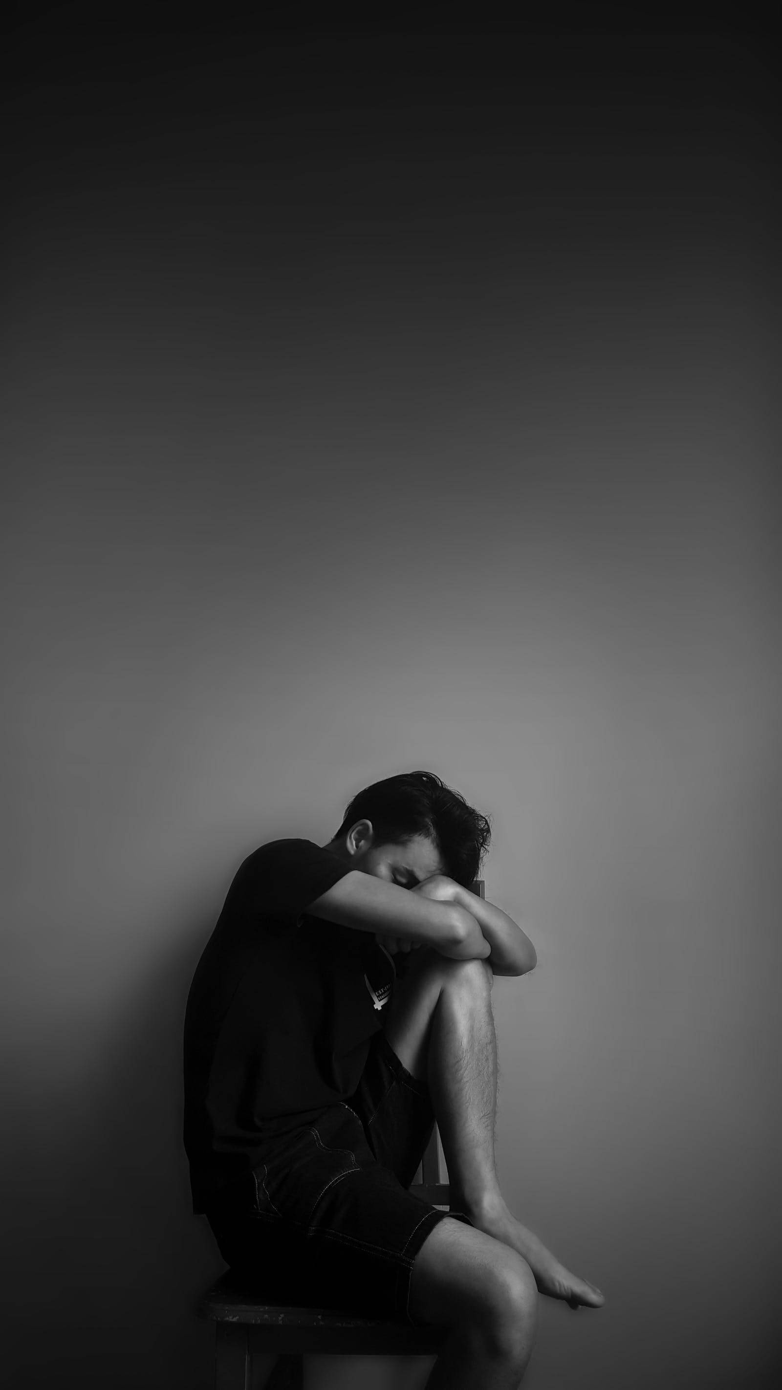 孤獨男子倚牆而坐 壁紙 — 一個人獨自坐在矮凳上，背靠素牆，低頭屈膝，雙手環抱膝蓋，一幅黑白靜默絕望的肖像。