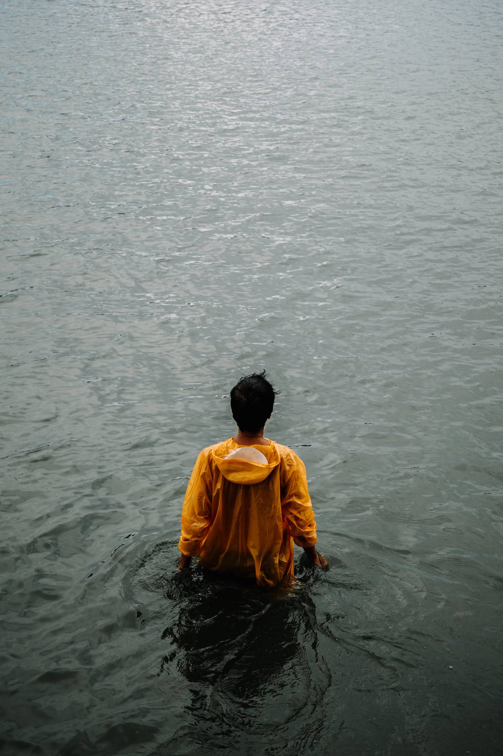 孤獨男子立於水中 壁紙 — 一個人穿著鮮黃色雨衣，腰部浸在平靜的灰色水中，從背後觀看。