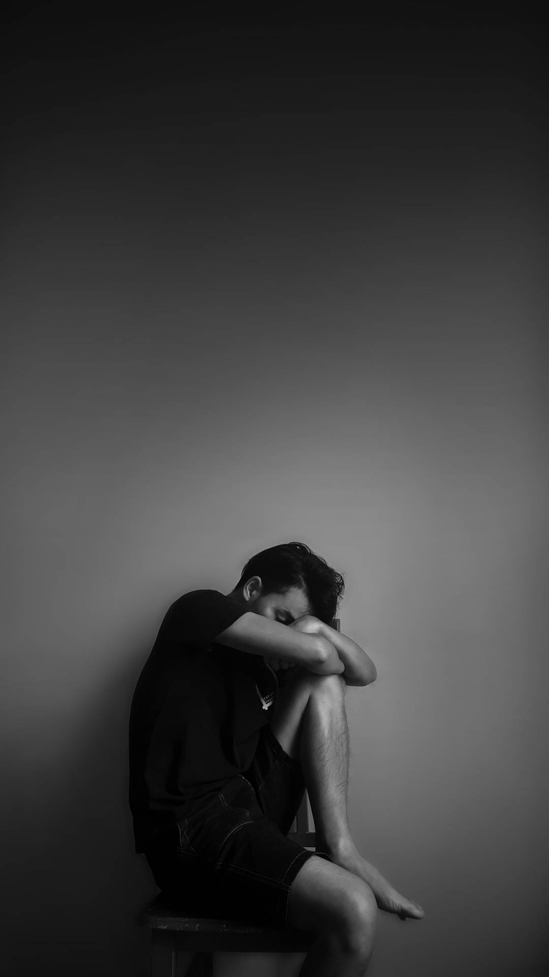 一個人獨自坐在凳子上，低著頭，在灰階房間中。