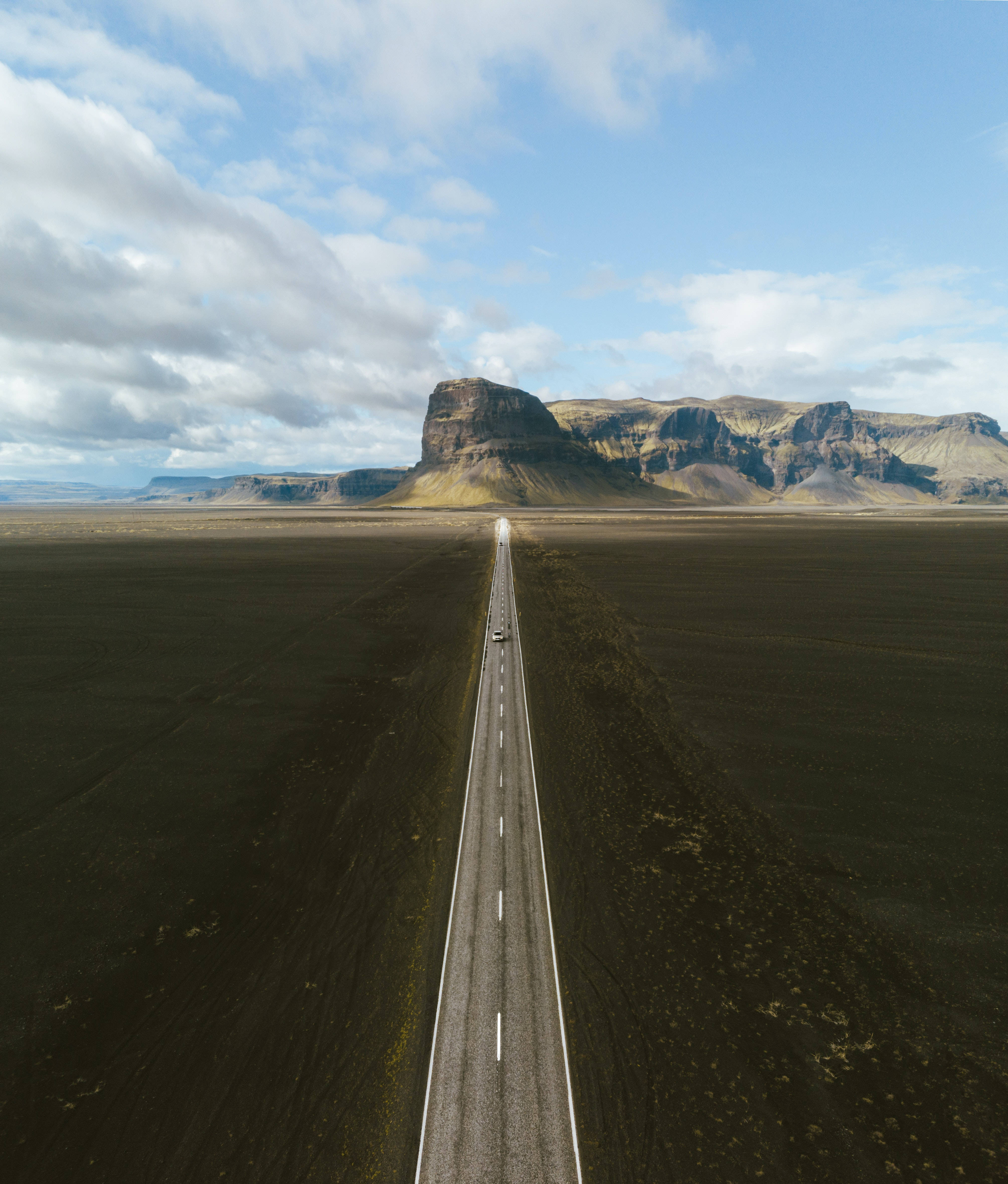 孤獨道路雲背景 壁紙 — 一輛孤獨的汽車沿著一條筆直的道路穿過黑色火山平原，駛向遠處崎嶇的山脈，天空部分多雲。