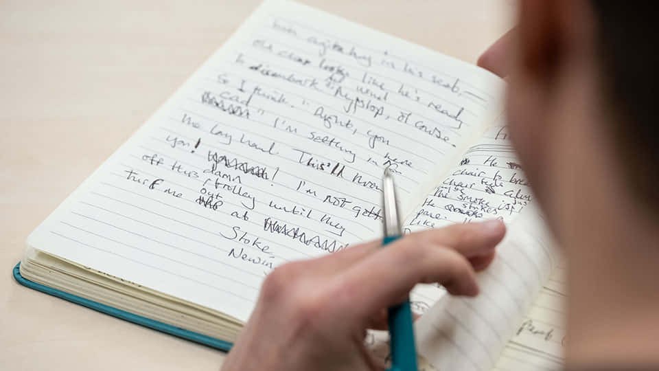 手用藍色筆喺木桌上嘅間線筆記本上寫字。