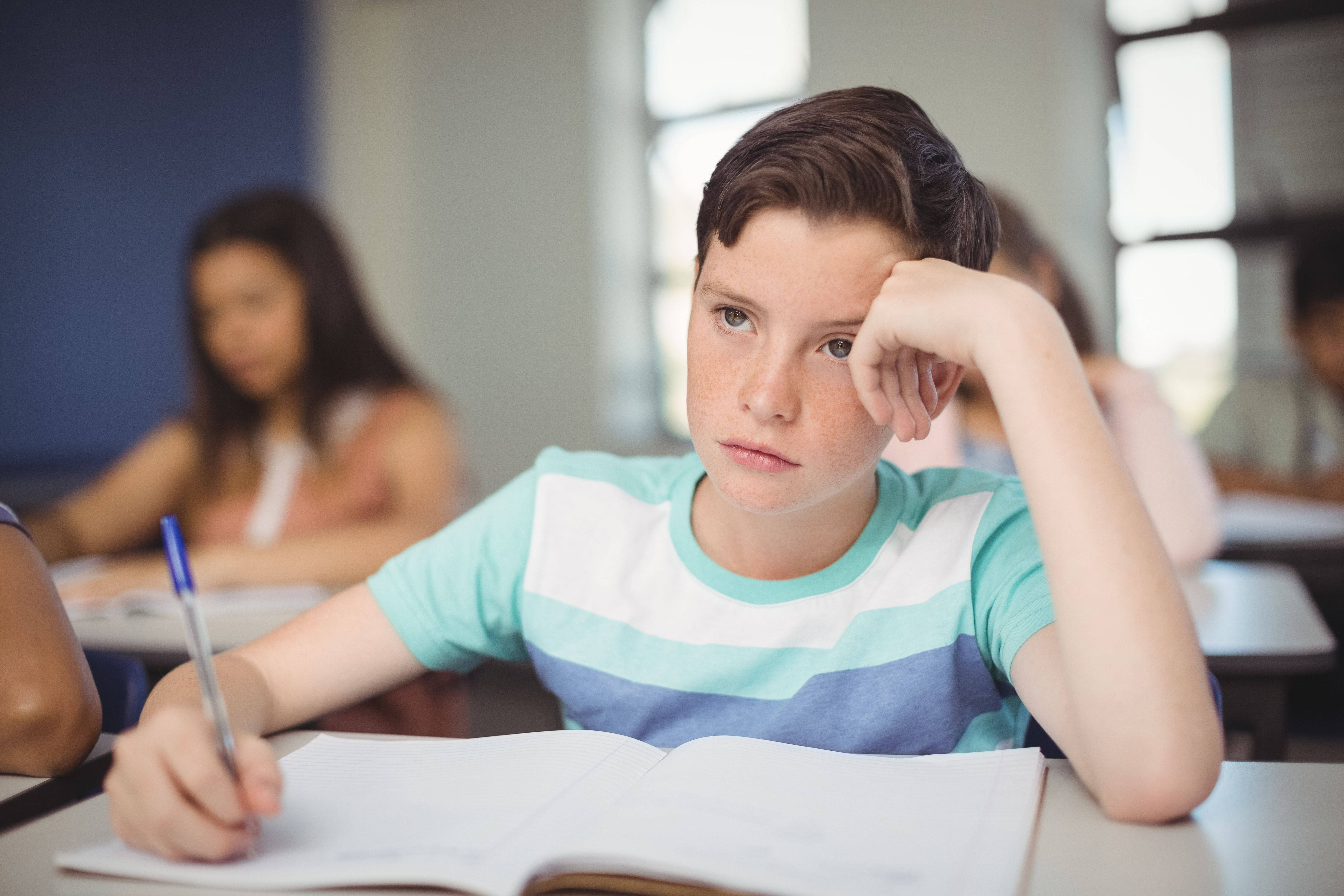 學習中的沉思 壁紙 — 一個無聊嘅青少年男仔將頭挨喺手上，同時喺課室入面揸住一支筆喺打開嘅筆記簿上面。