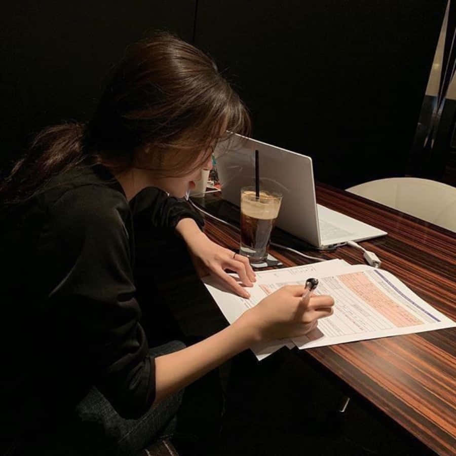 學習女孩與咖啡和筆記本電腦圖像 壁紙 — 一個人在昏暗的房間裡，俯身在一張木桌上，在筆記本電腦和冰咖啡旁邊寫字。