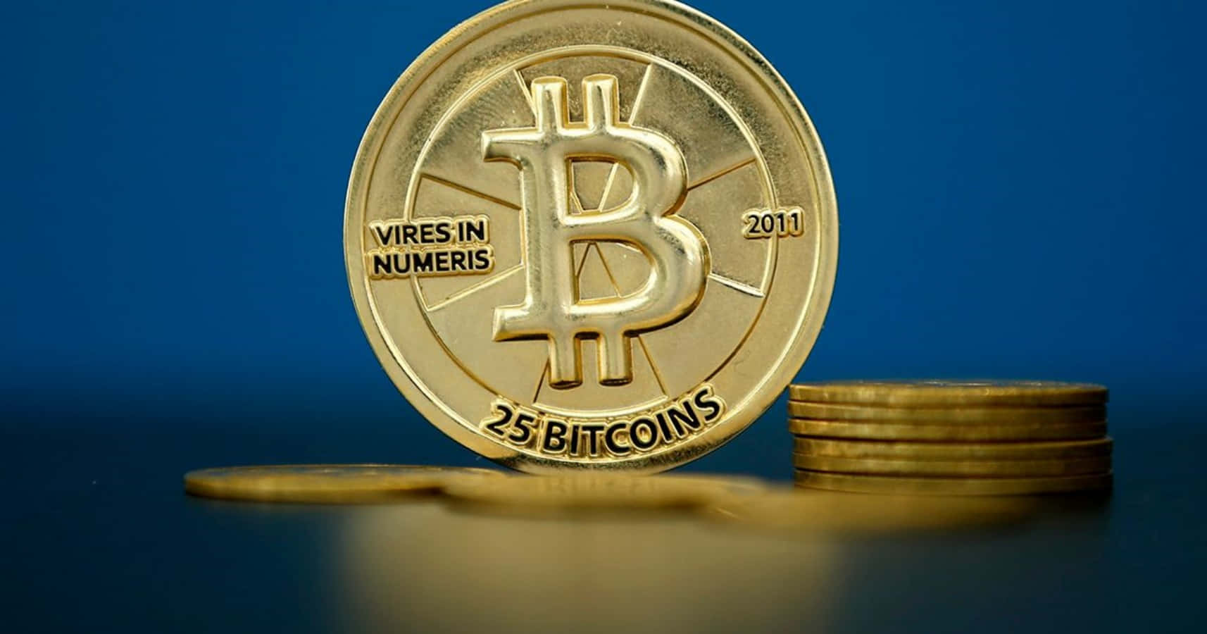 學習比特幣，體驗財務自由 壁紙 — 一枚閃閃發光的金色比特幣硬幣，上面刻有「25 BITCOINS」和「2011」，直立在一堆硬幣旁邊，背景是深藍色。