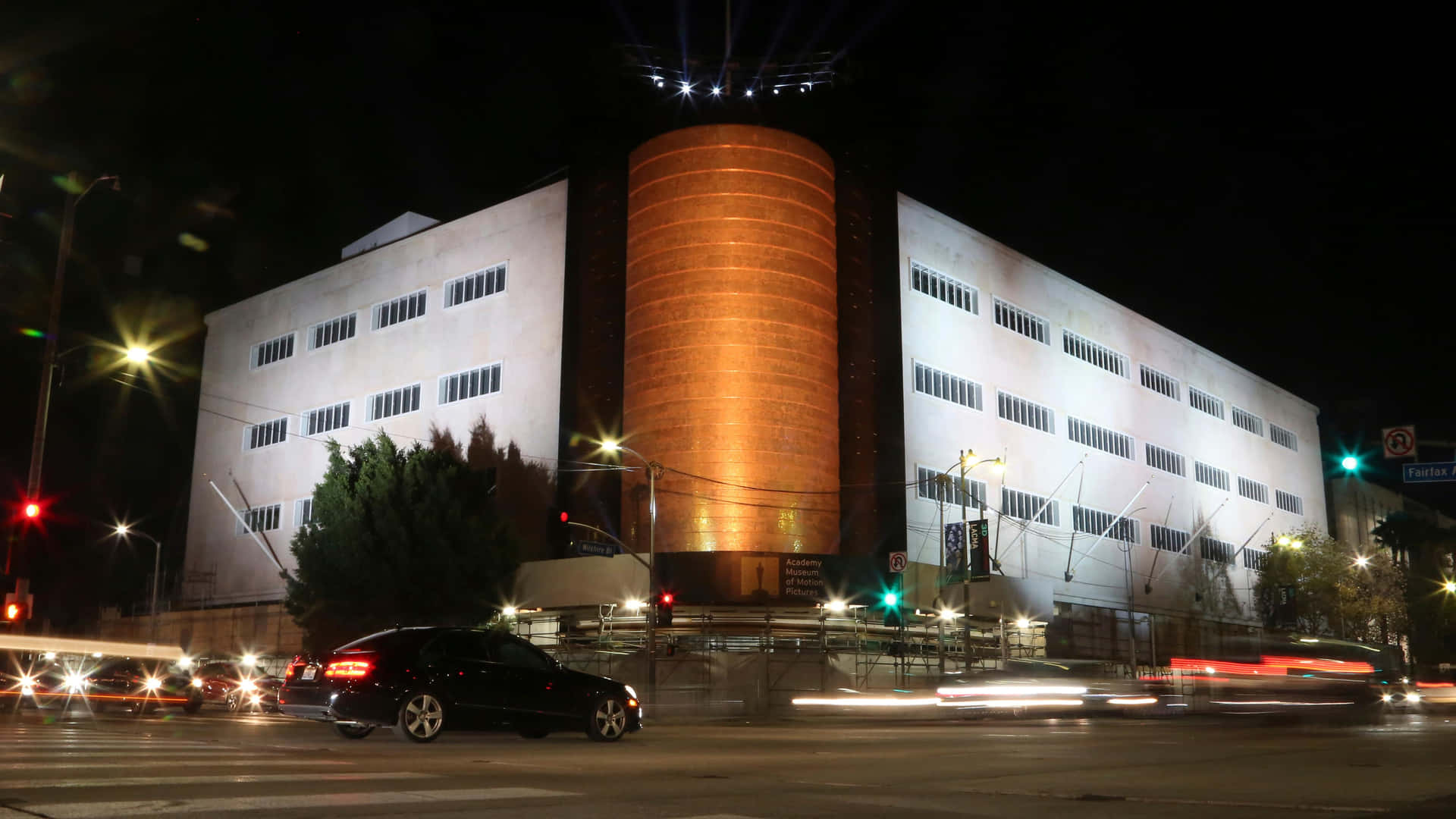 學院電影博物館夜間美景 壁紙 — 一座現代建築，帶有發光的橙色圓柱形塔樓，在夜晚矗立，汽車帶著光跡疾馳而過。