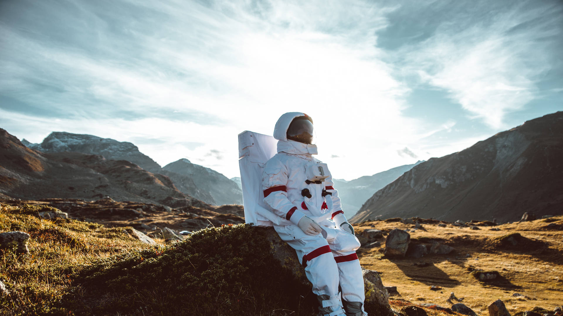 身穿白色太空衣的太空人坐在岩石地形上，背景是日落時分的山脈。