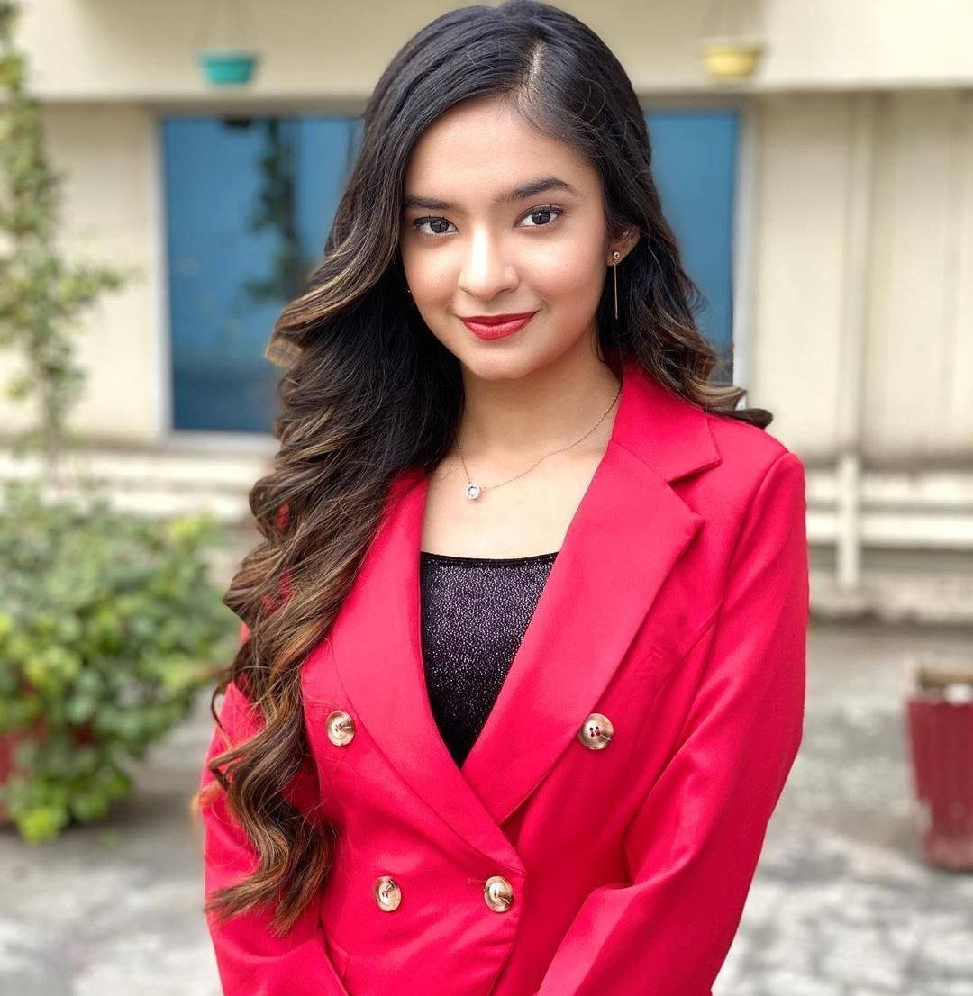 穿紅色西裝外套的年輕女性在戶外靠近建築物和植物微笑。