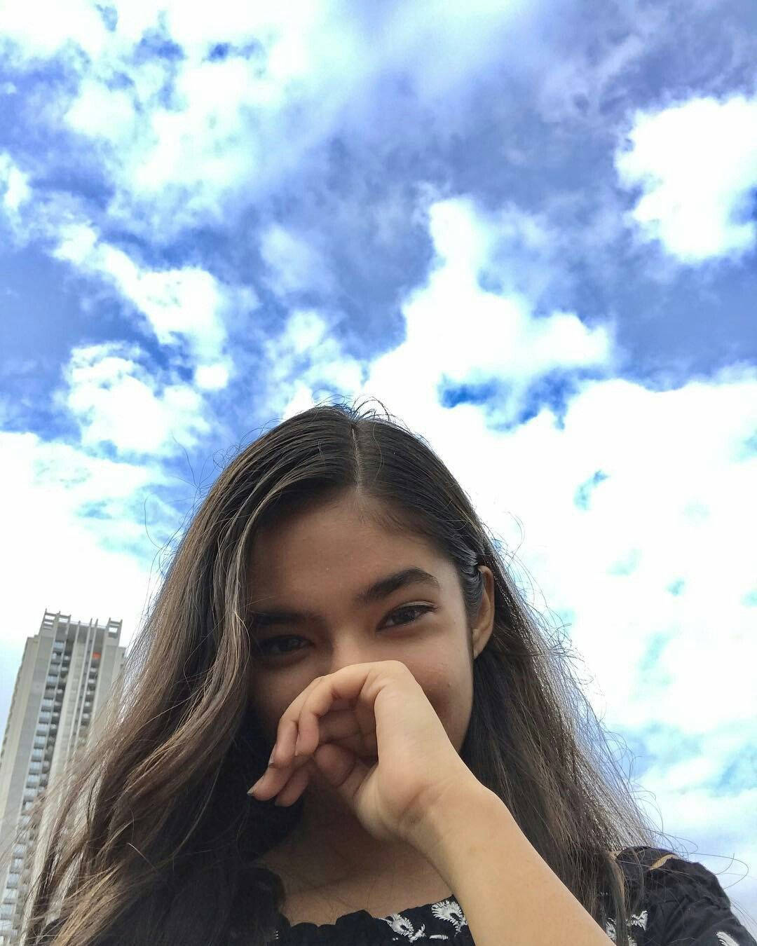 年輕女性在戶外多雲的天空下，背景有建築物，頑皮地捂住嘴巴。