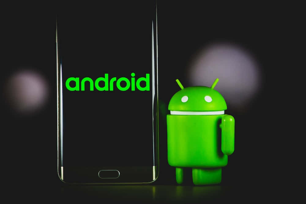 安卓標誌 壁紙 — 一個綠色Android吉祥物公仔站在一部顯示Android標誌的智能手機旁邊，背景為深色模糊。