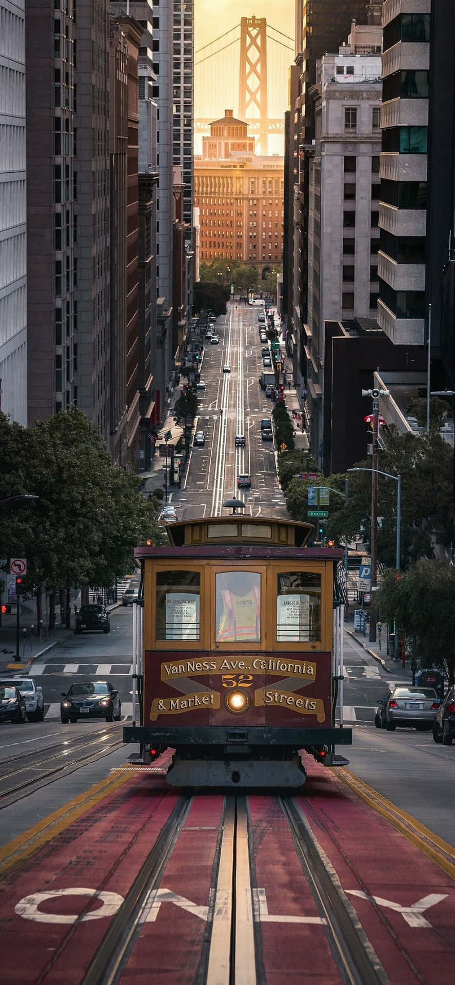 安卓聖弗朗西斯科纜車背景 壁紙 — 一輛經典的纜車沿著陽光明媚的三藩市街道滑行，兩旁是高聳的摩天大樓，遠處是標誌性的金門大橋。