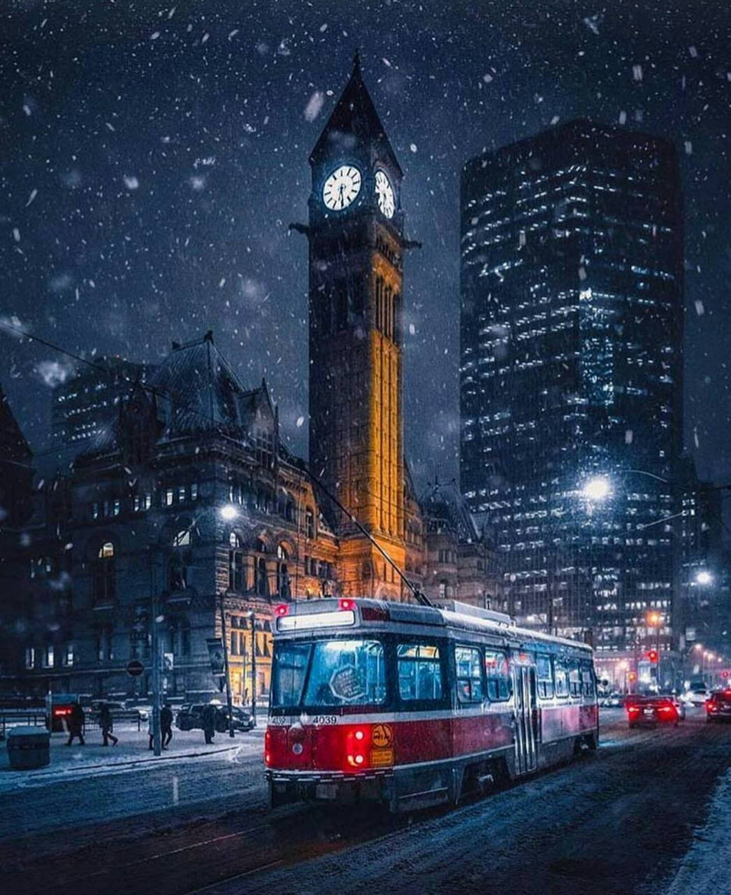 安大略鐘樓 壁紙 — 一輛紅色電車在雪夜中滑行，城市中歷史悠久的鐘樓和現代摩天大樓在飄落的雪花下閃閃發光。