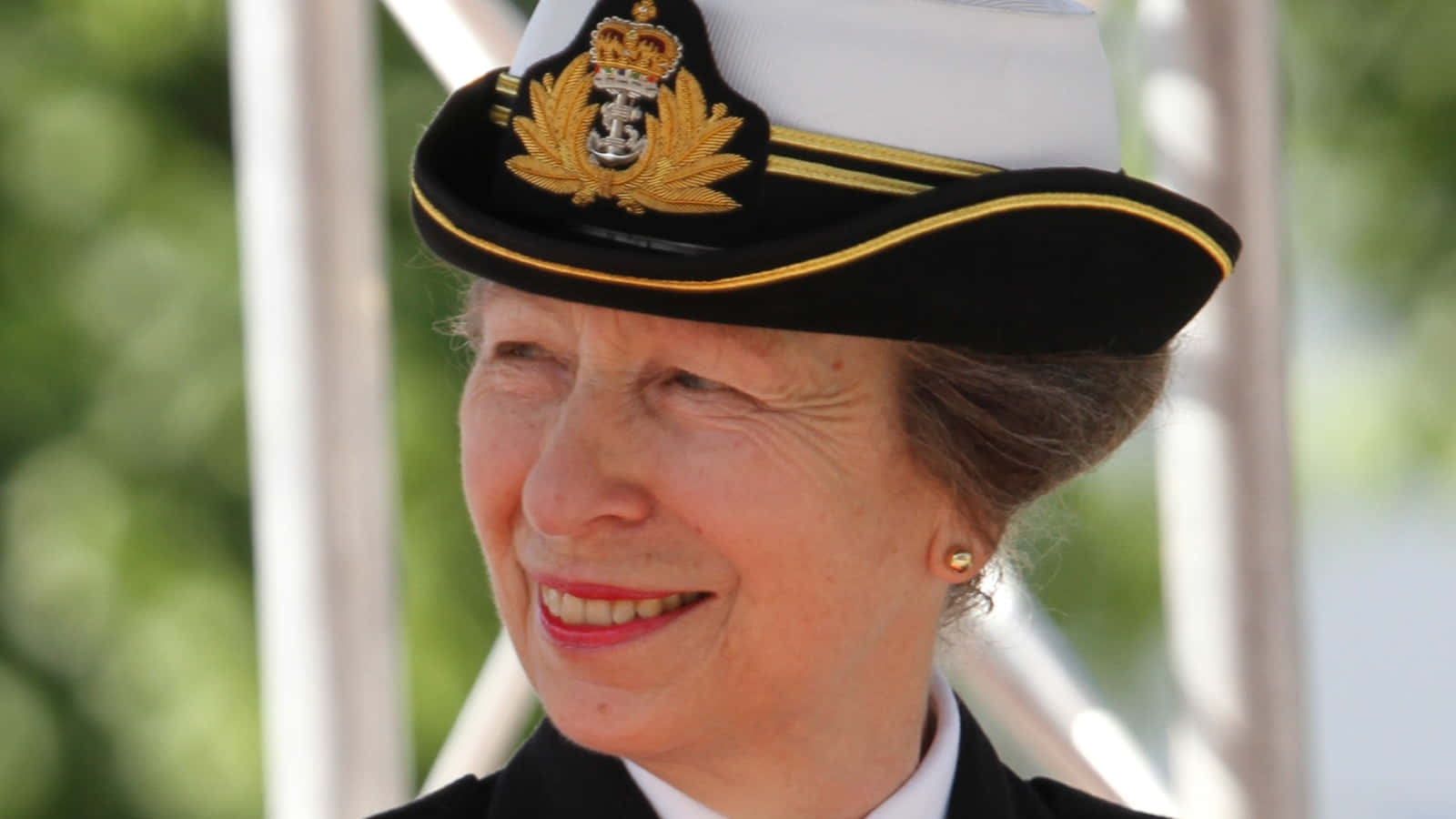 安妮公主佩戴皇家海軍帽 壁紙 — 一位戴著海軍軍官帽的老年女性，在模糊的綠色和白色背景前溫暖地微笑。