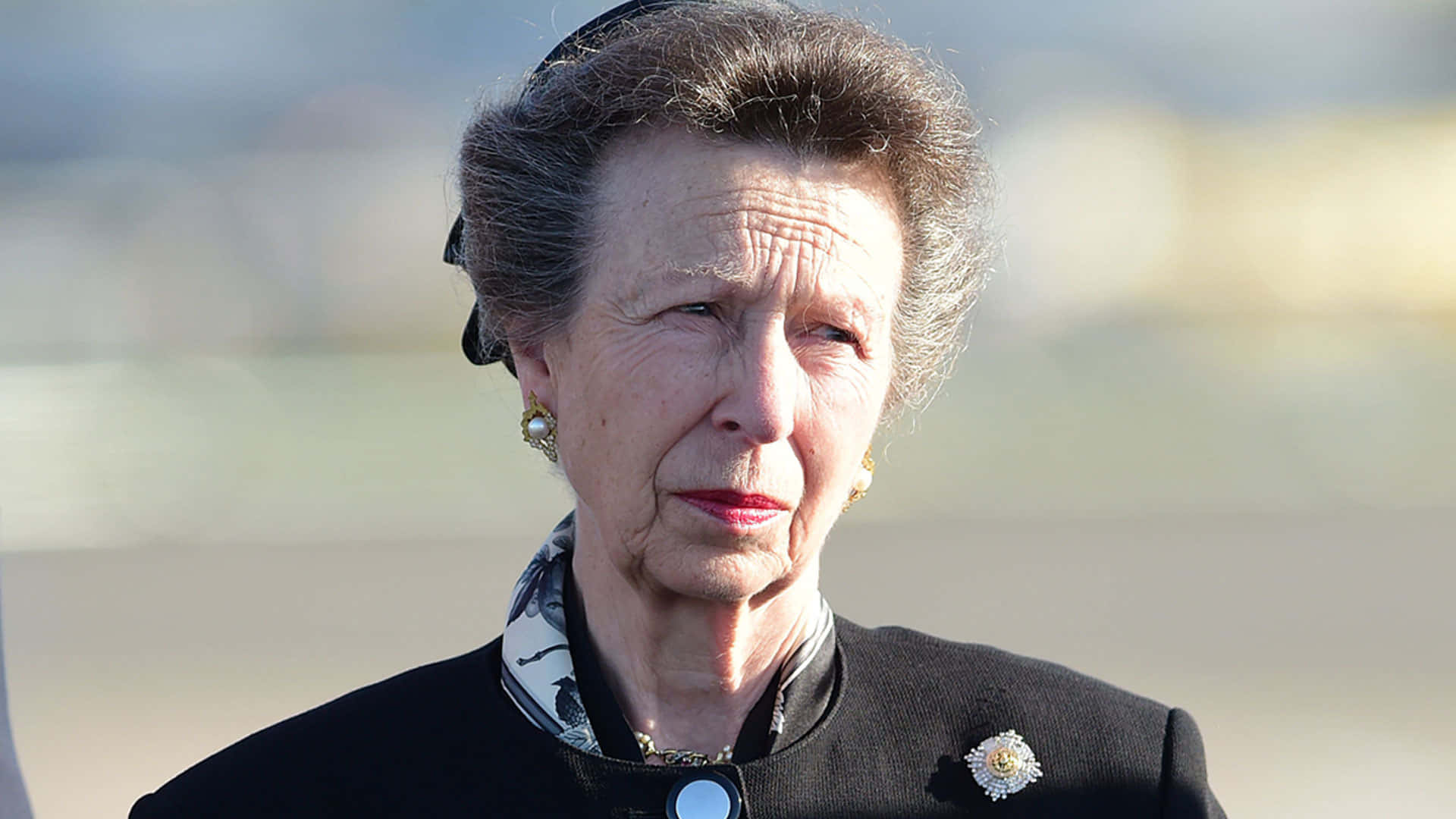 安妮公主哀傷神情 壁紙 — 灰髮戴珍珠耳環的老年女性，身穿黑色外套，別著花卉胸針，背景為模糊的戶外景色。