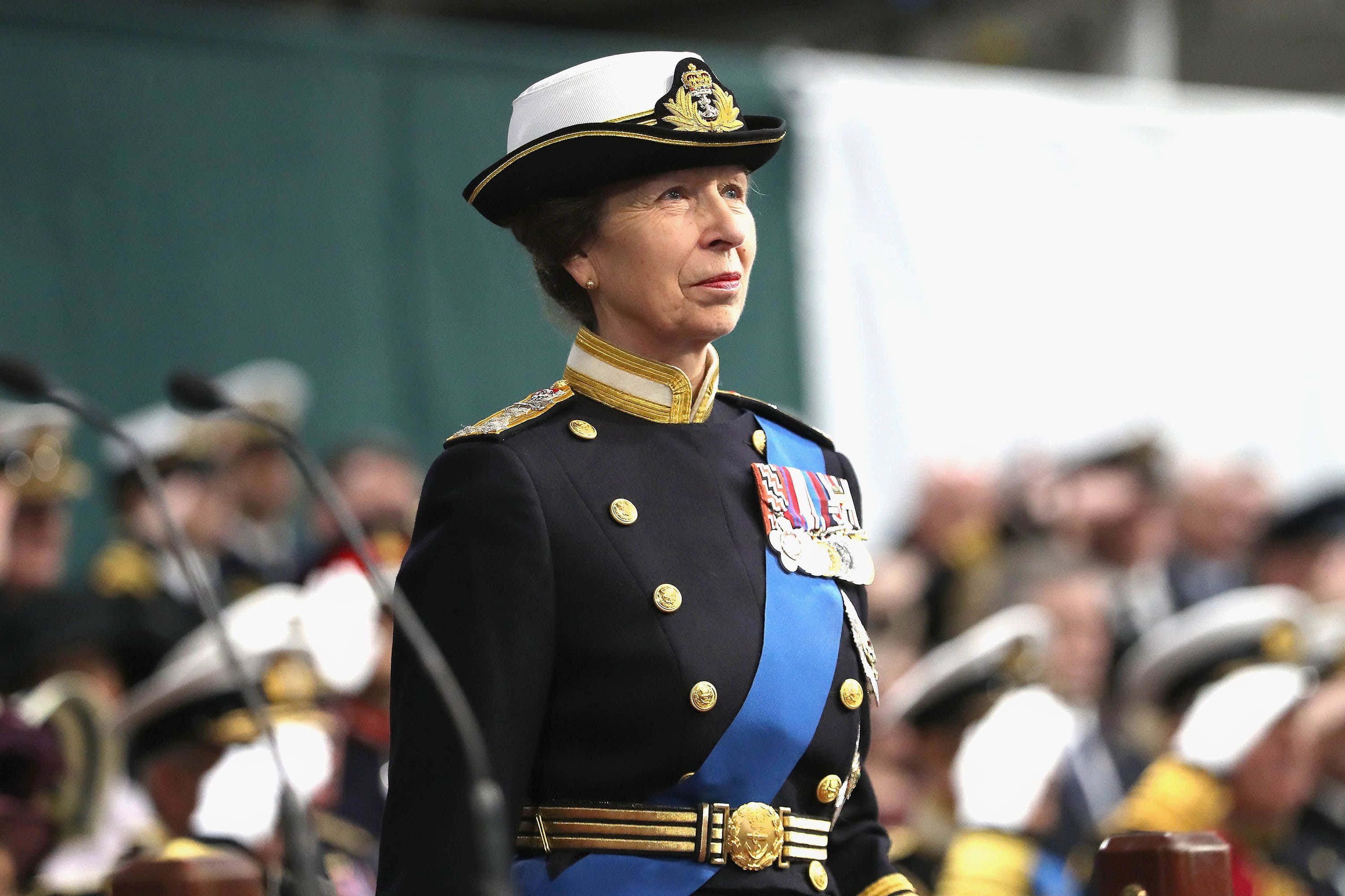 安妮公主身著皇家軍裝 壁紙 — 一名身穿正式海軍制服、佩戴勳章和金色飾邊的女性，在麥克風和模糊的與會者背景下，莊嚴地站在典禮活動中。